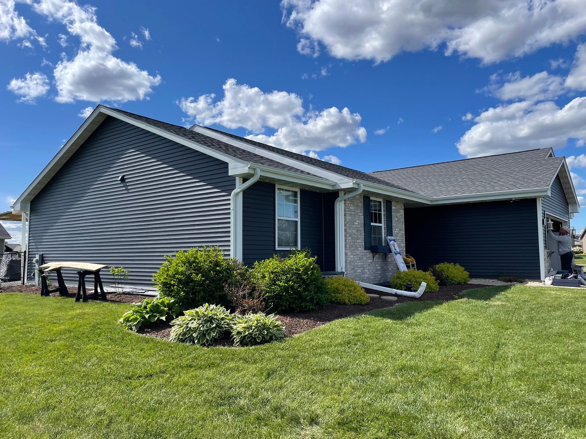 Blue-sided house with a dark gray roof on a sunny day. Green lawn and landscaping in the foreground.
