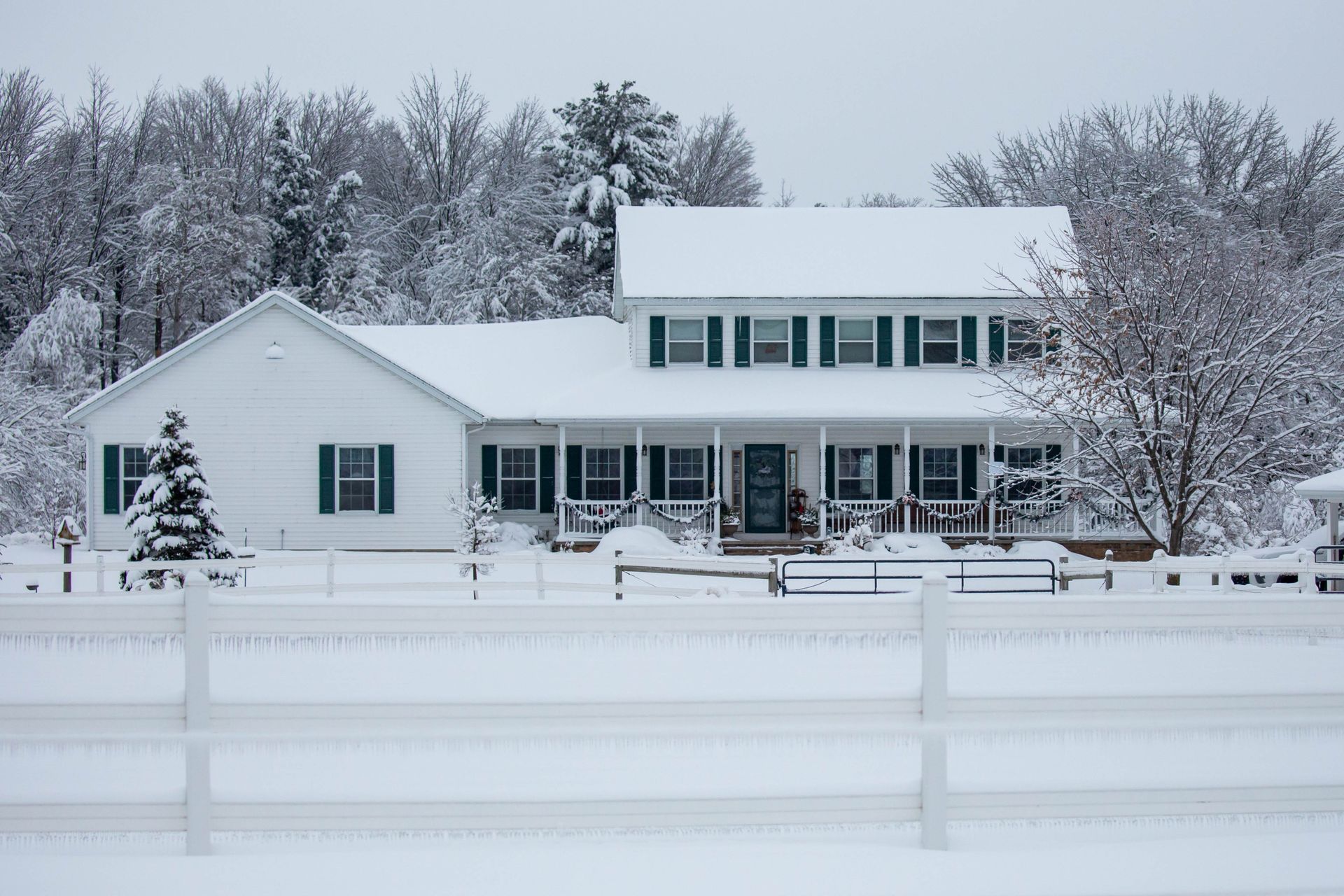 White two-story house with green shutters and snow-covered roof, surrounded by snow and trees. A white fence is in front.