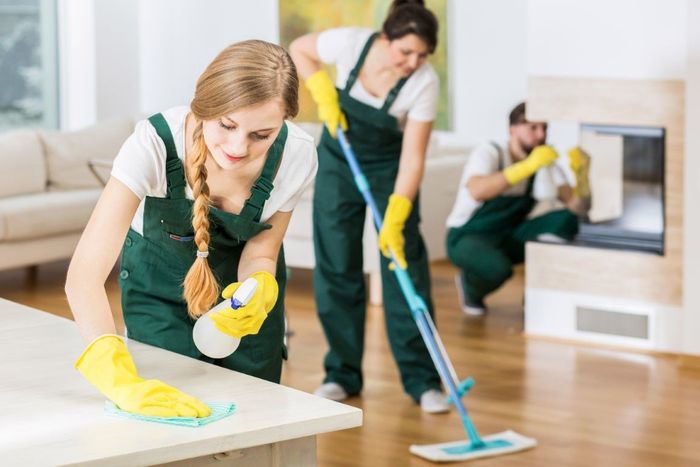 Three cleaning professionals in green jumpsuits, yellow gloves, are cleaning a bright room; one wipes a table, one mops a floor, and one cleans a fireplace.