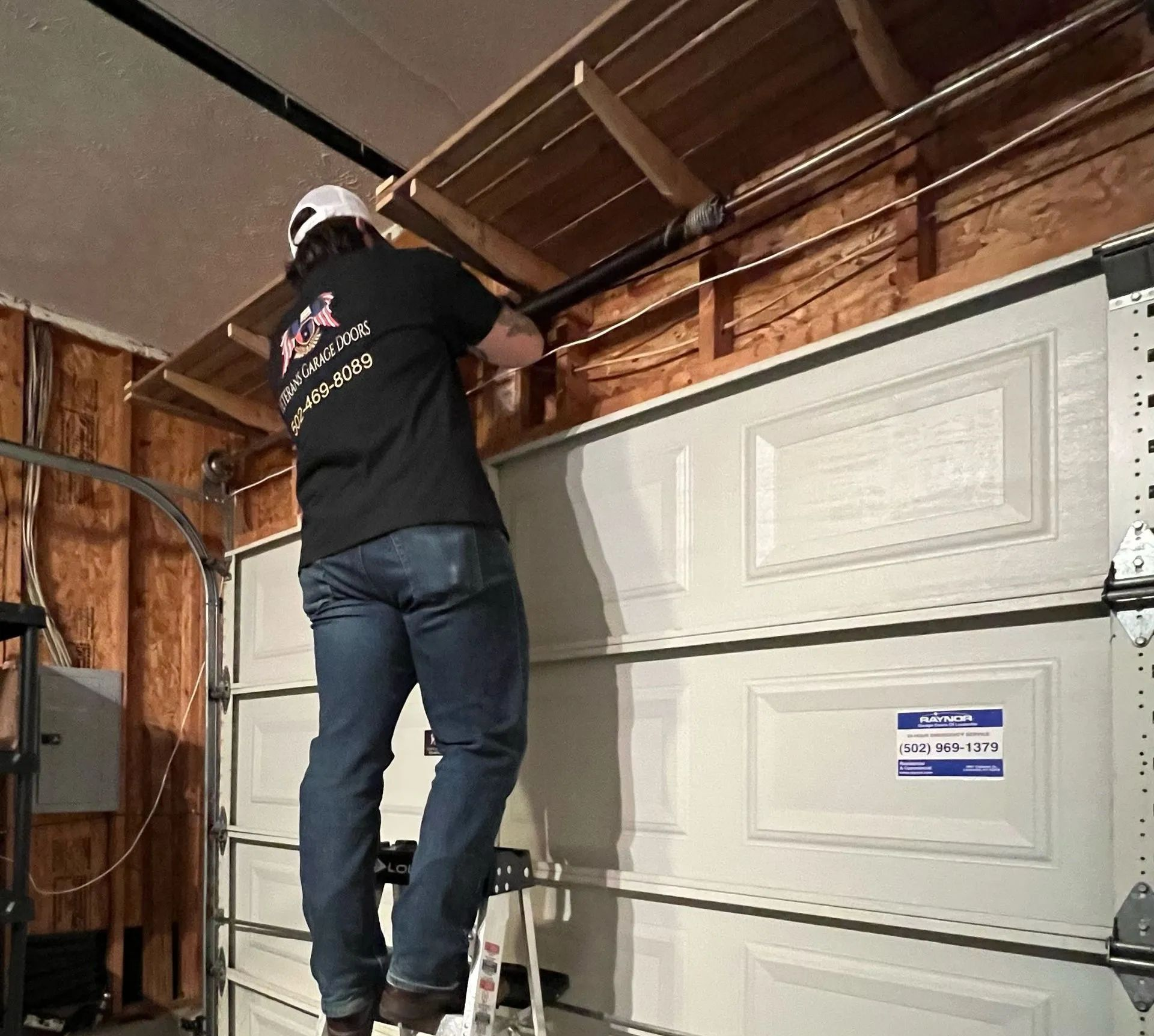 A technician standing on a ladder while repairing the garage door opener mechanism mounted to the ceiling.