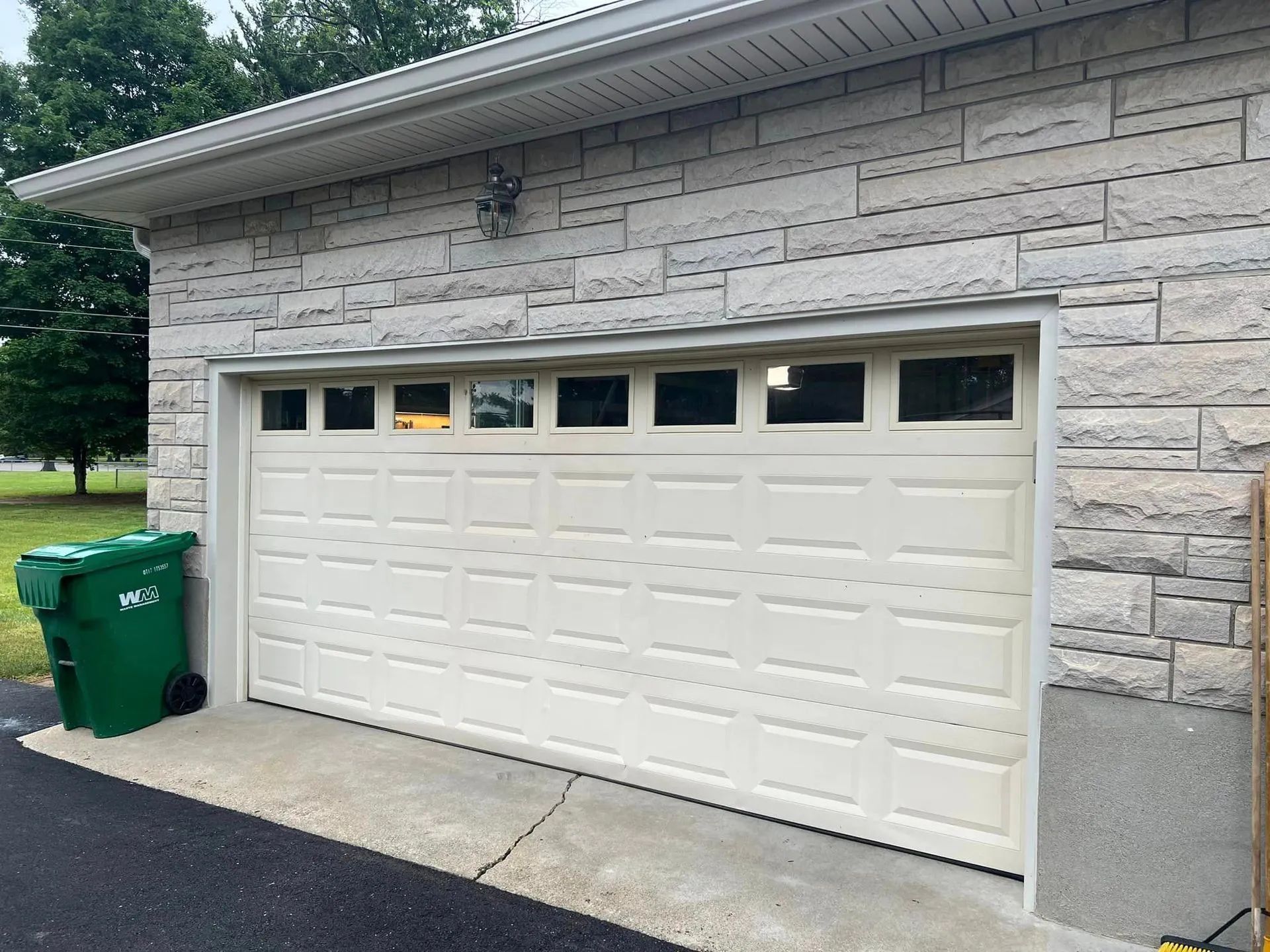 White garage door with window panels, in front of a light brick building. A green trash bin is to the left.
