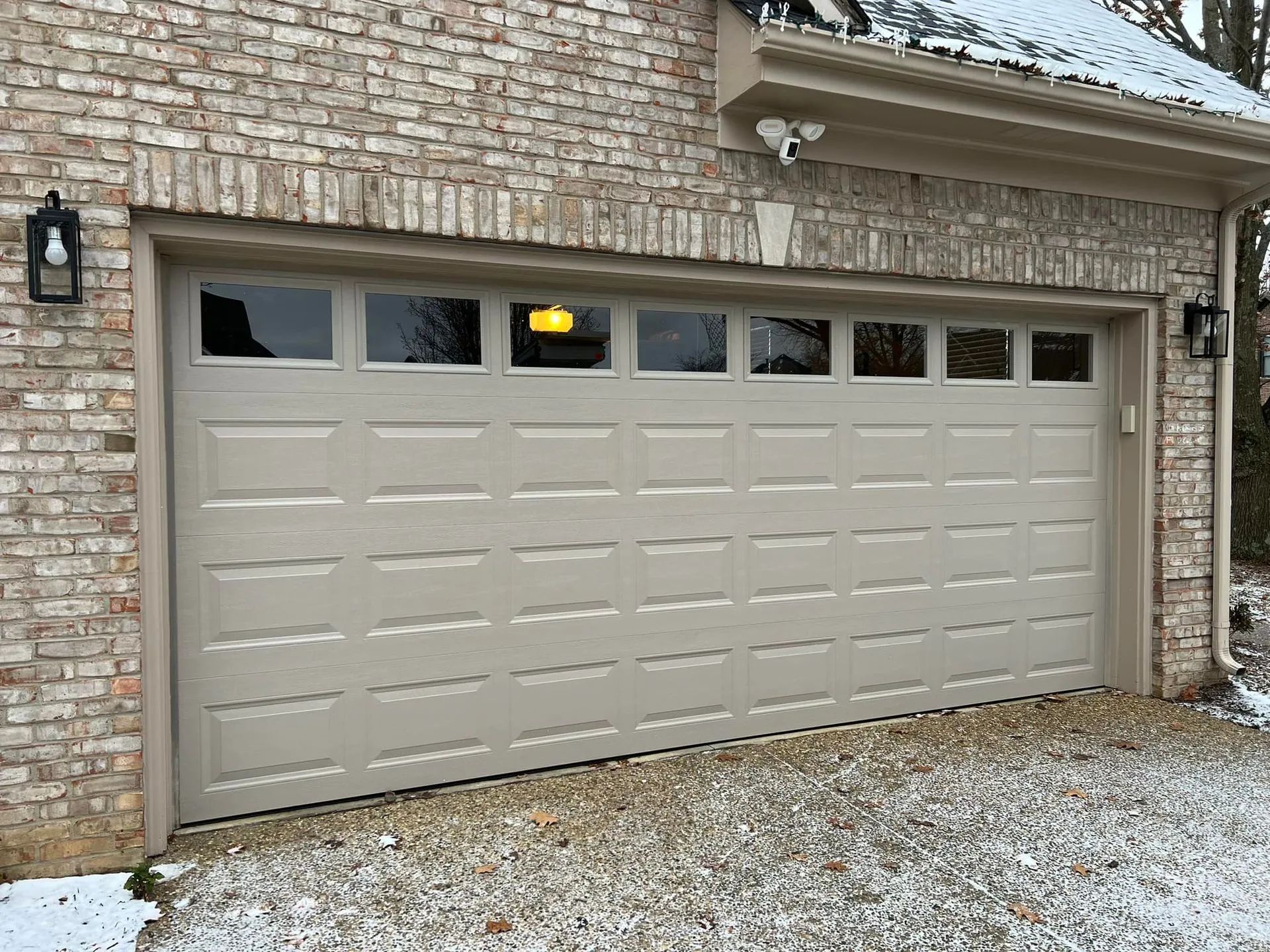 Tan garage door on a brick house with windows and black lights, lightly covered in snow.