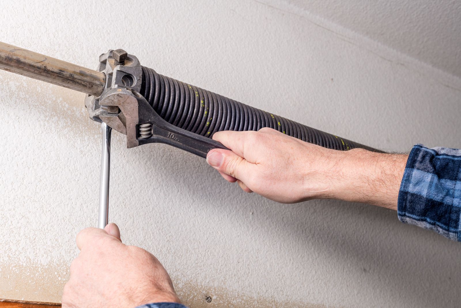 Person using a wrench to adjust a garage door spring, against a white wall.