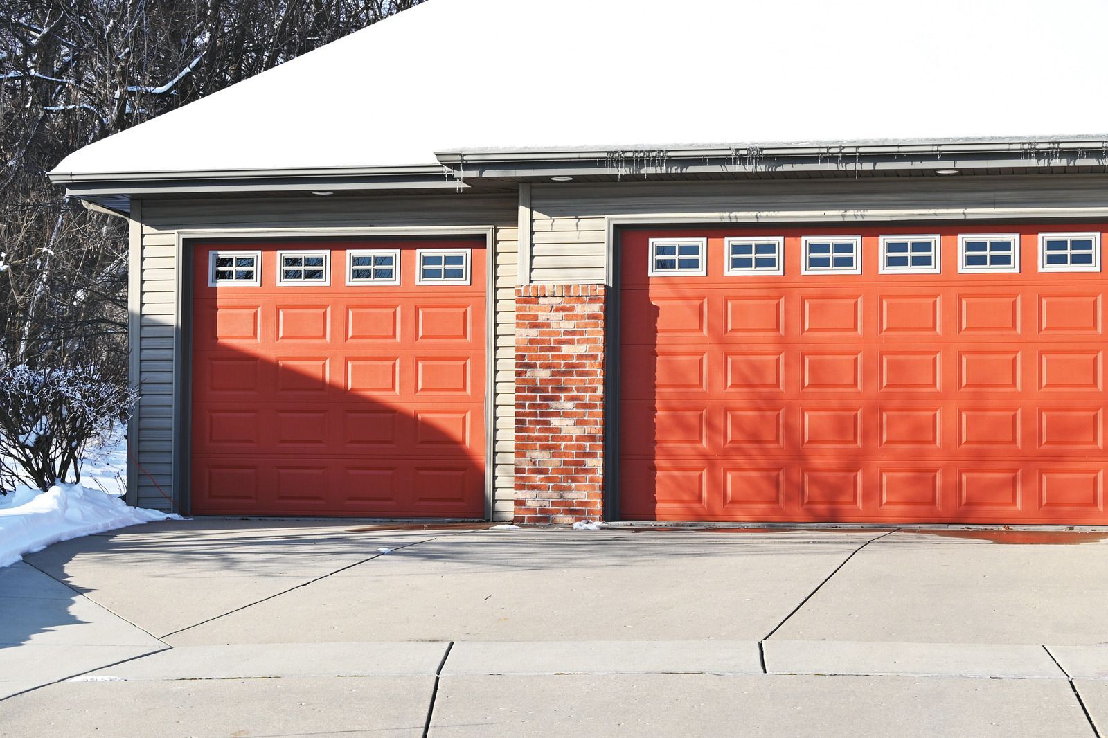 Two orange garage doors with square windows, snow on the roof, and a brick pillar in between.