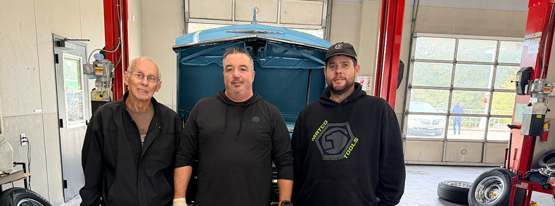 Three men standing in a garage beside a blue car hood, with tools and equipment in the background | Motorvation Service Center