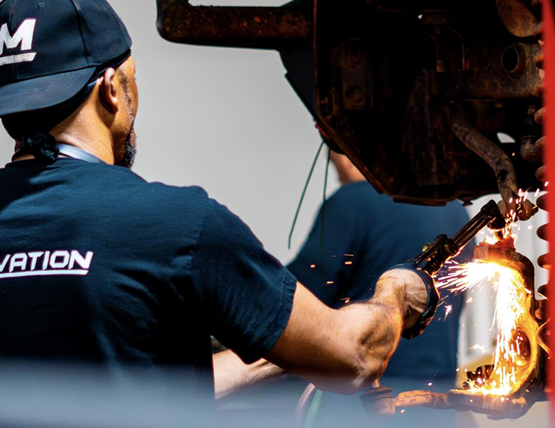 Worker welding metal on a truck chassis, sparks flying in a dark workshop | Motorvation Service Center