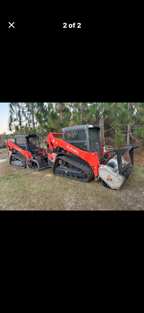 Two red Kubota track loaders parked on a grassy field, one equipped with a forestry mulcher attachment.