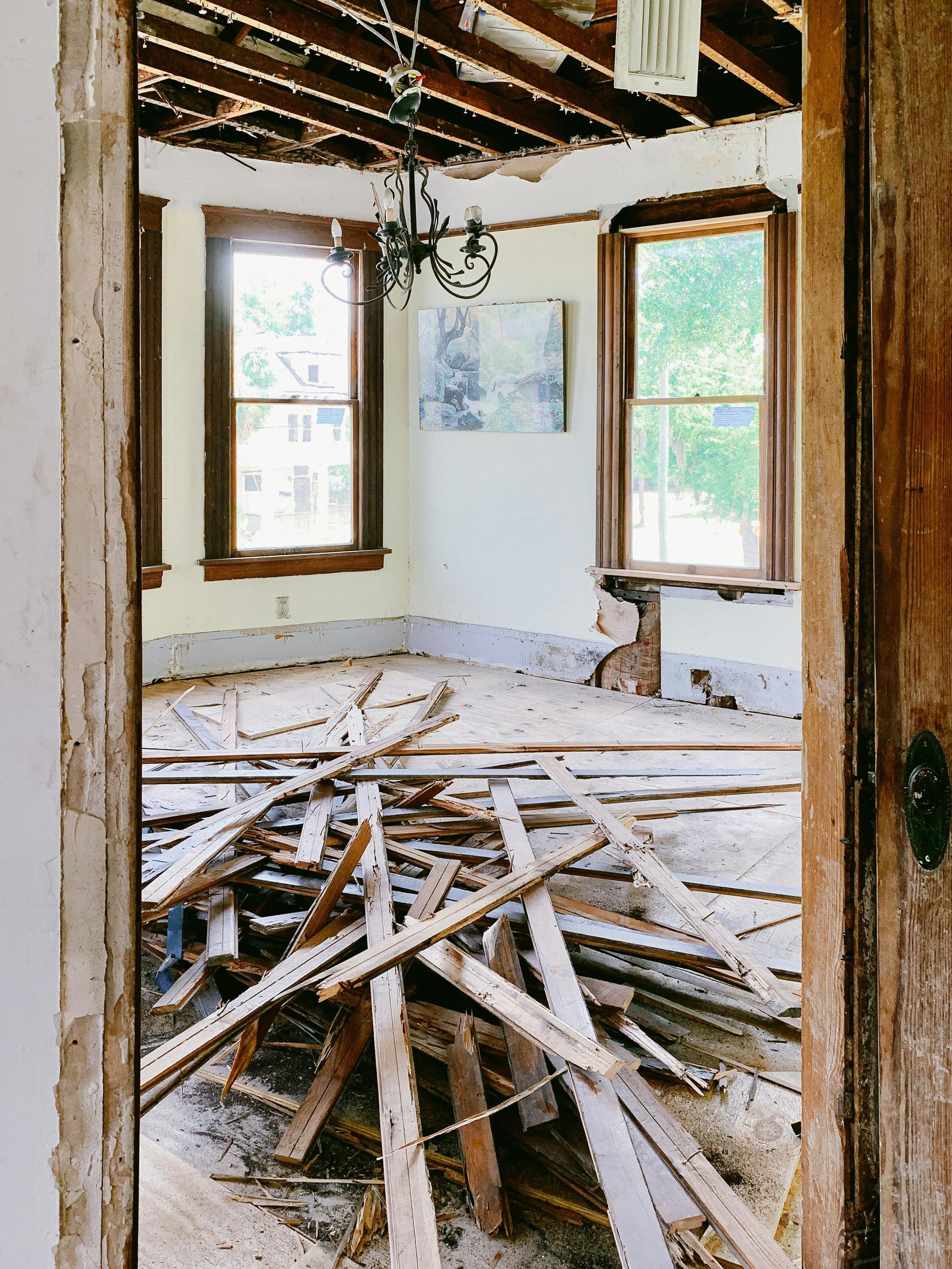 Room under renovation, debris-covered floor, exposed ceiling beams, windows, chandelier, and doorway.