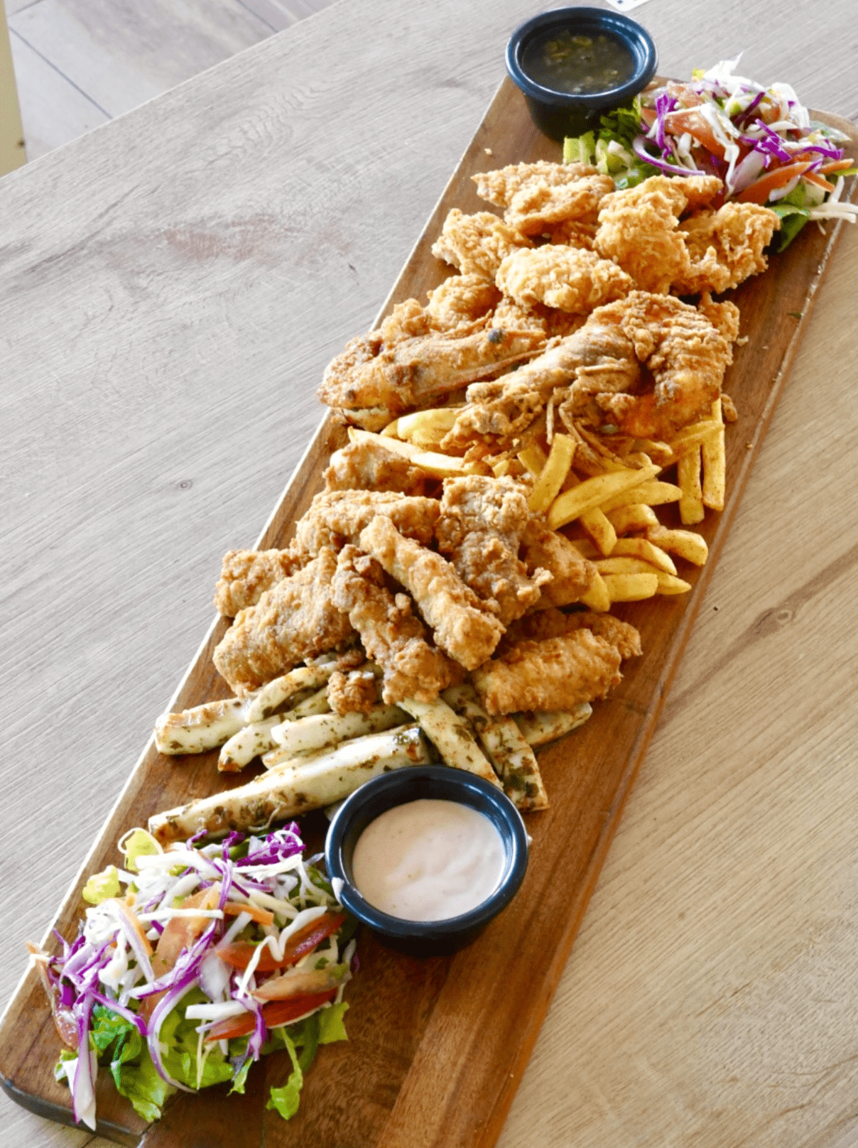 Una tabla de cortar de madera larga llena de comida frita y verduras.