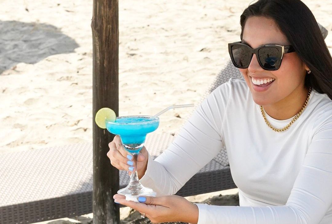 Una mujer con gafas de sol sostiene una margarita azul en la playa.