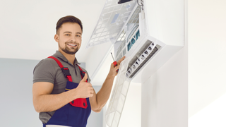 A man is standing on a ladder fixing an air conditioner and giving a thumbs up.