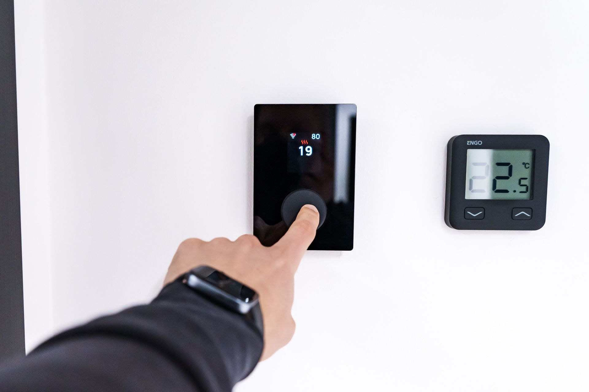 Hand pressing a black thermostat or control panel on a white wall beside a digital temperature display