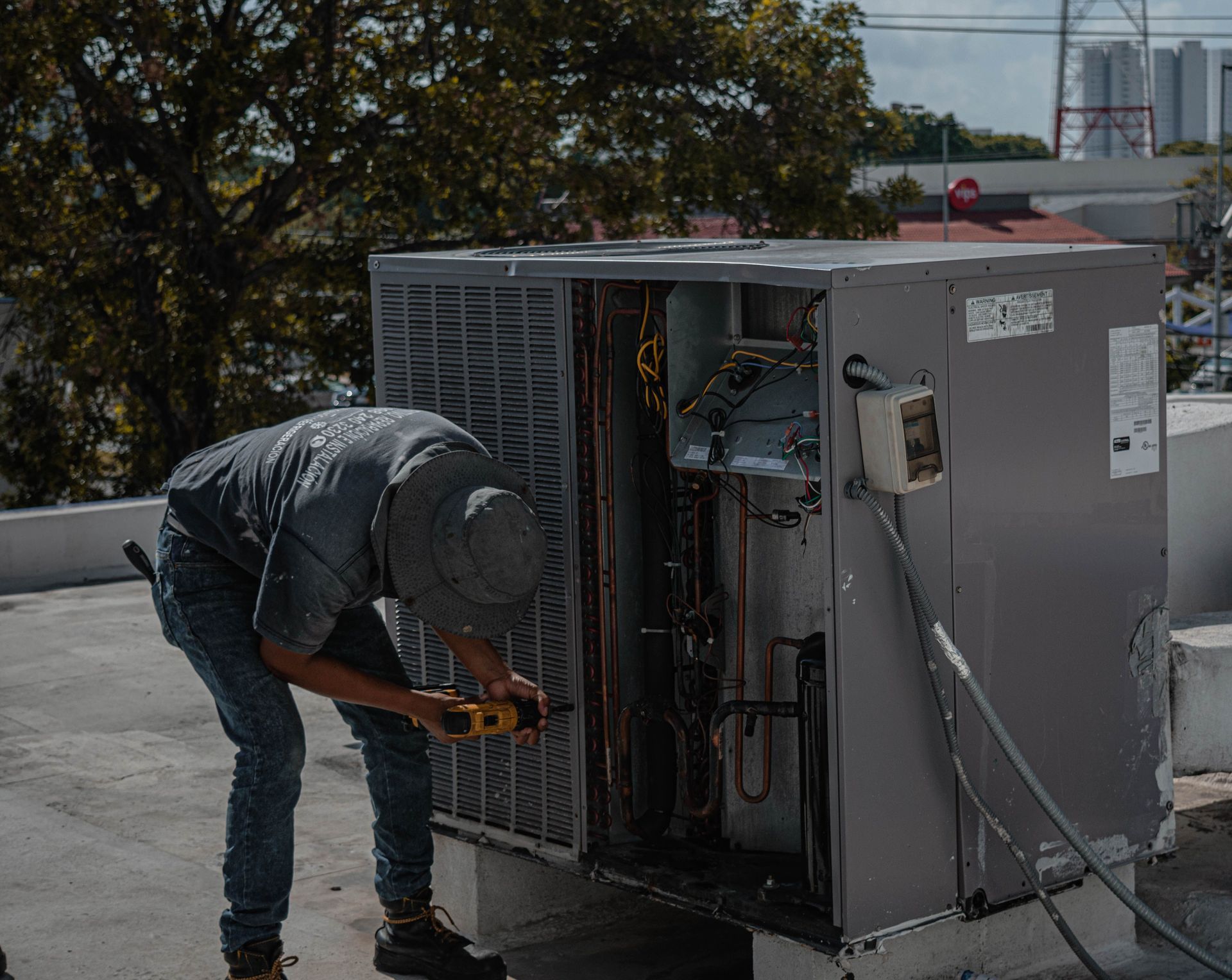 Technician crouches beside an open rooftop HVAC unit, inspecting wiring with tools.