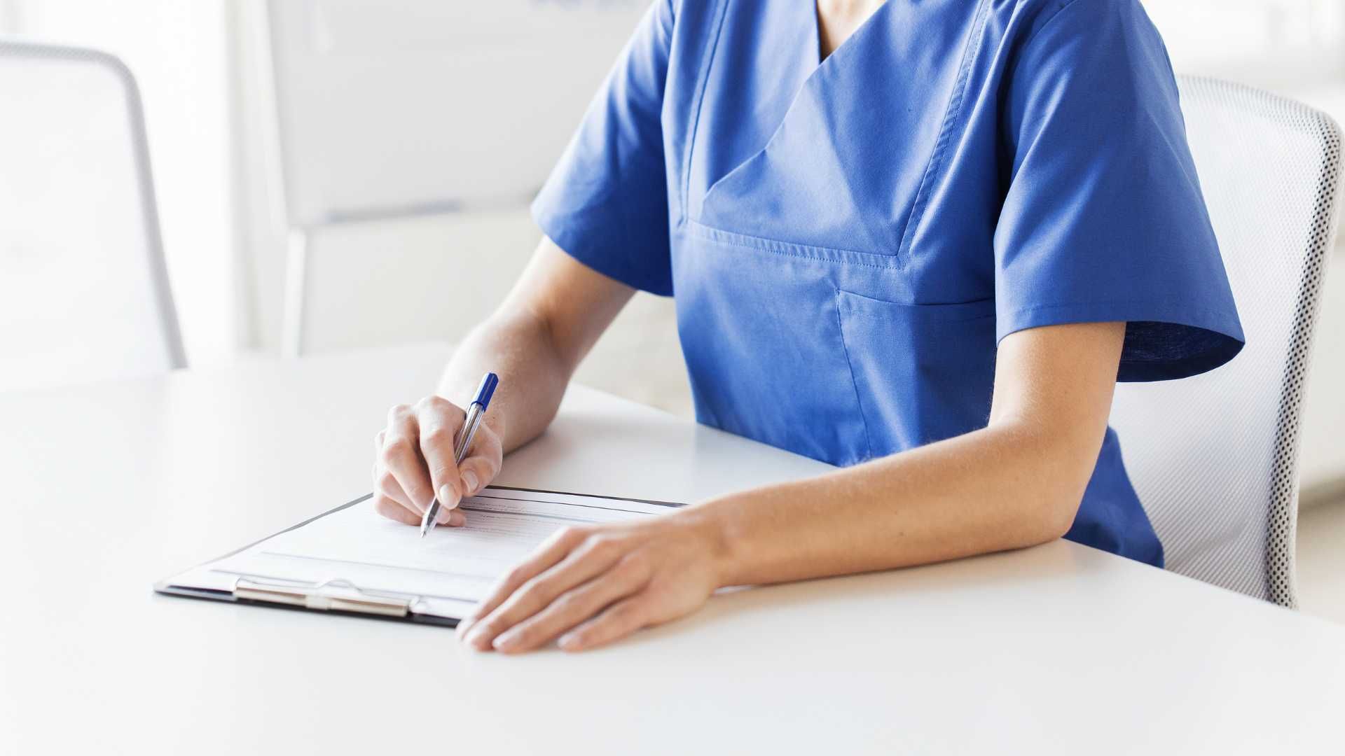 A nurse is sitting at a table writing on a clipboard.