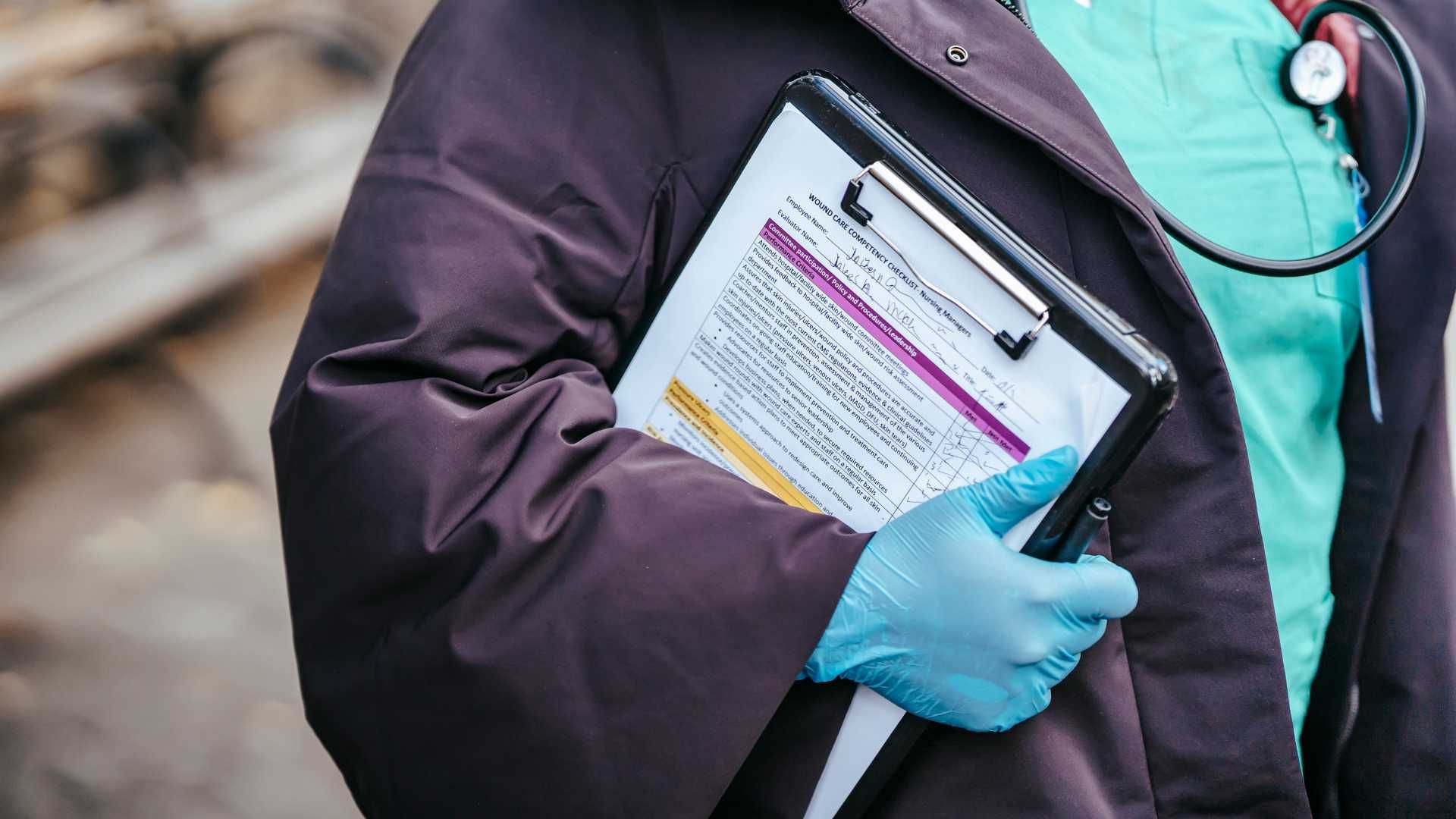 A doctor wearing blue gloves and a stethoscope is holding a clipboard.