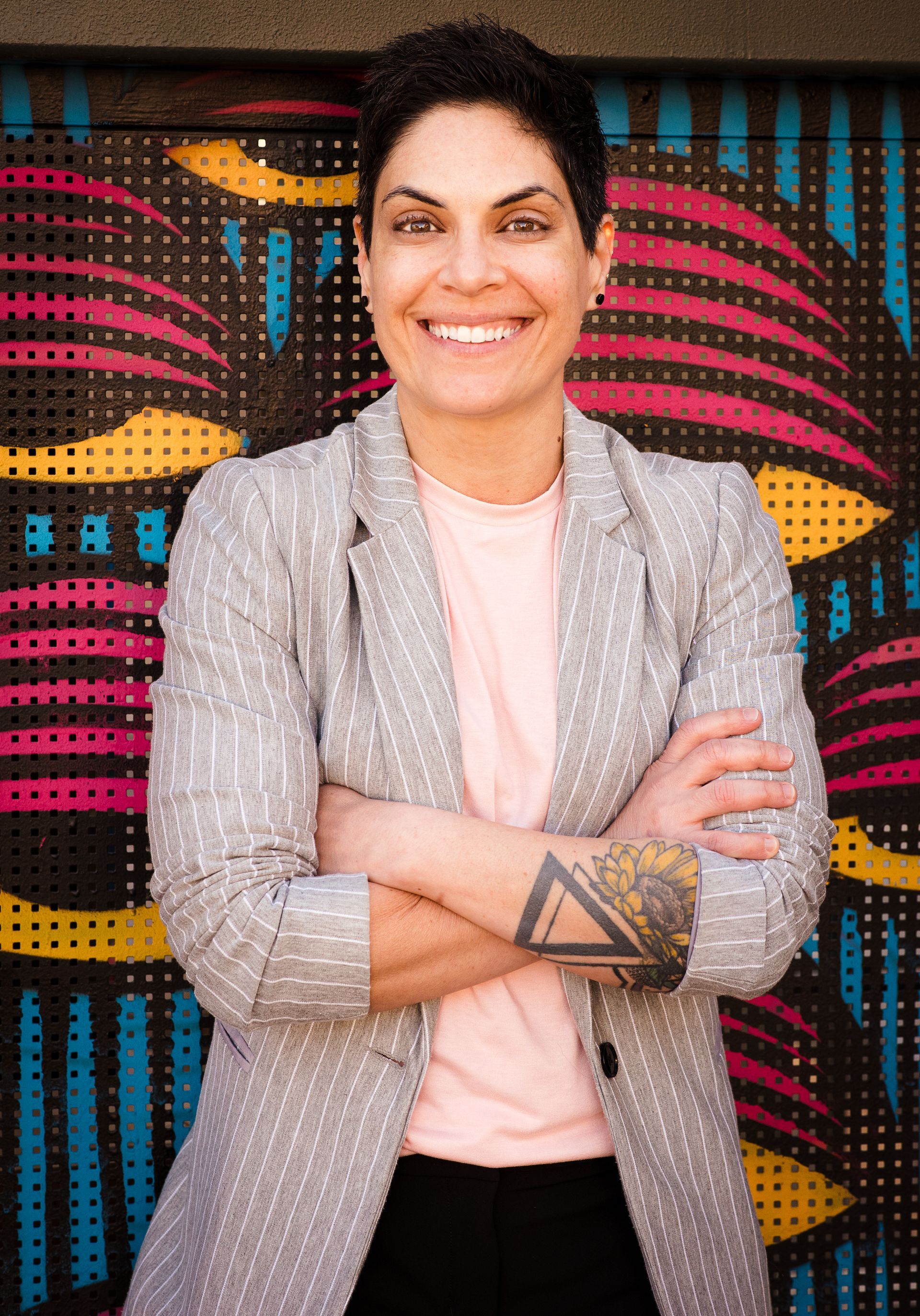 A woman with a tattoo on her arm is standing with her arms crossed in front of a colorful wall.