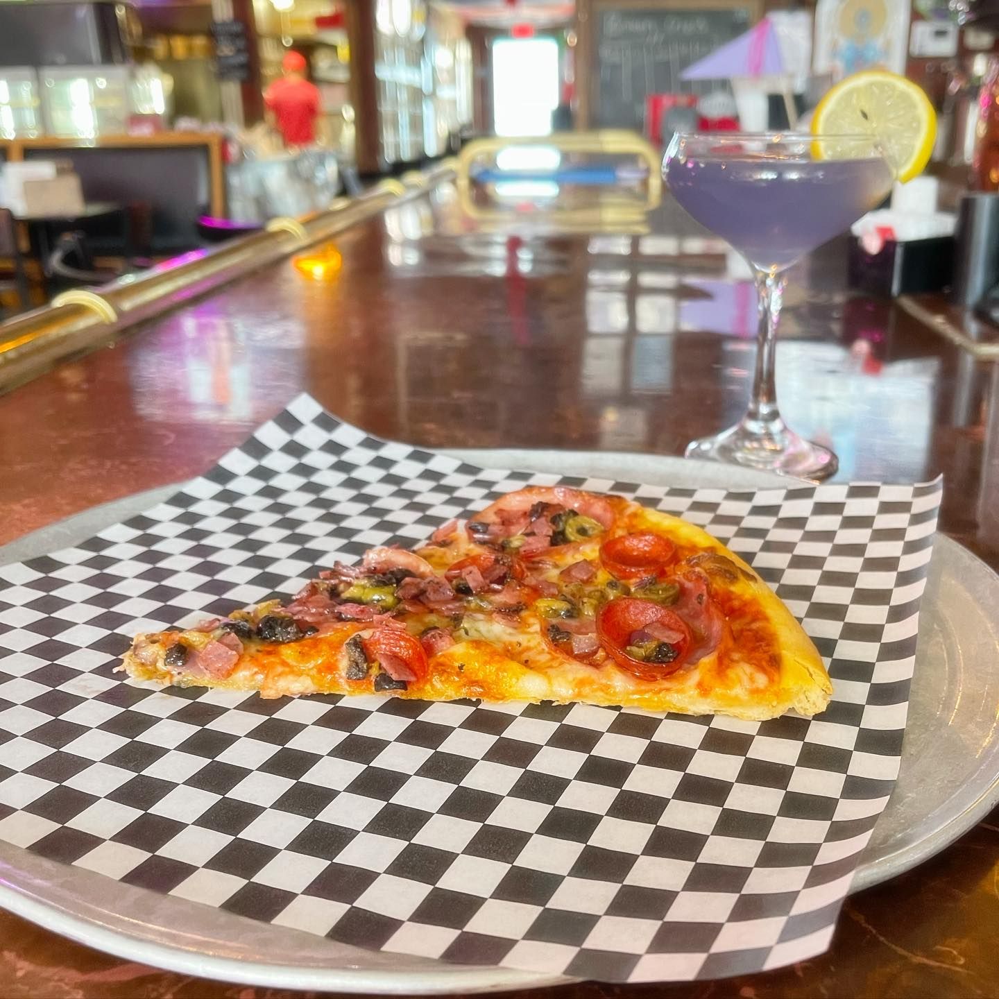 A slice of pizza on a checkered paper next to a martini glass