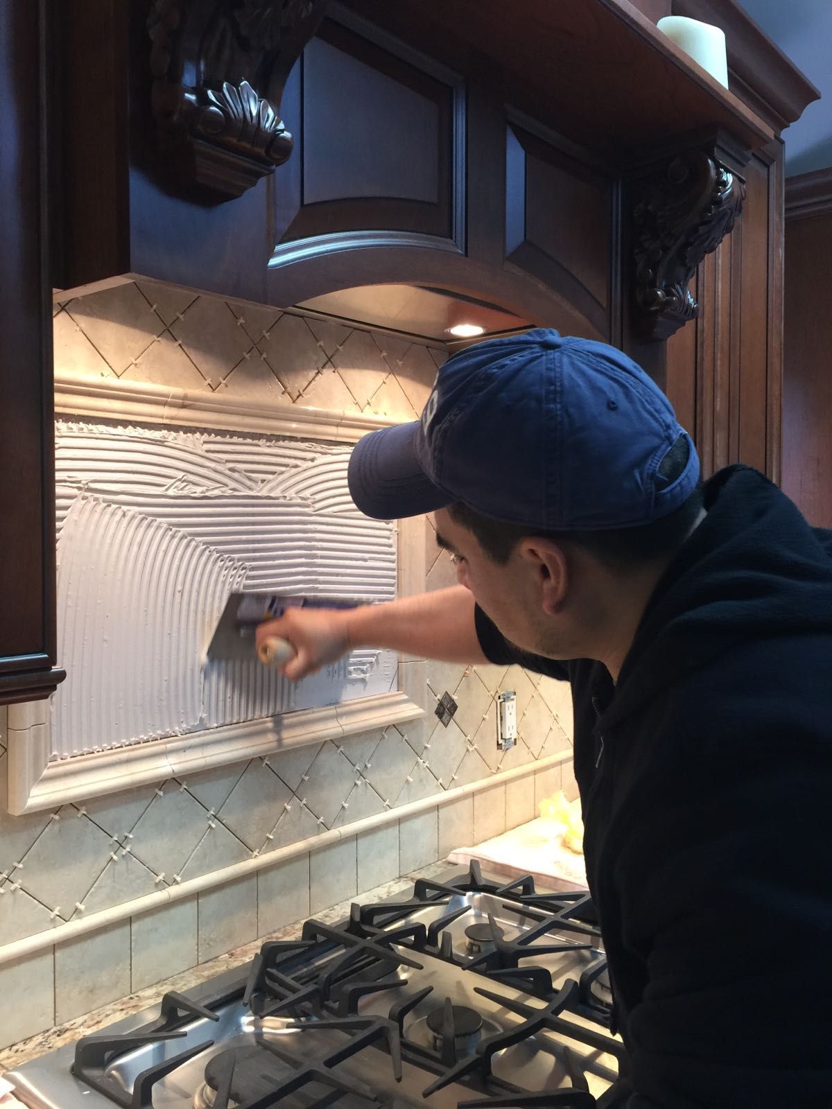 A man in a blue hat is working on a tile wall in a kitchen.