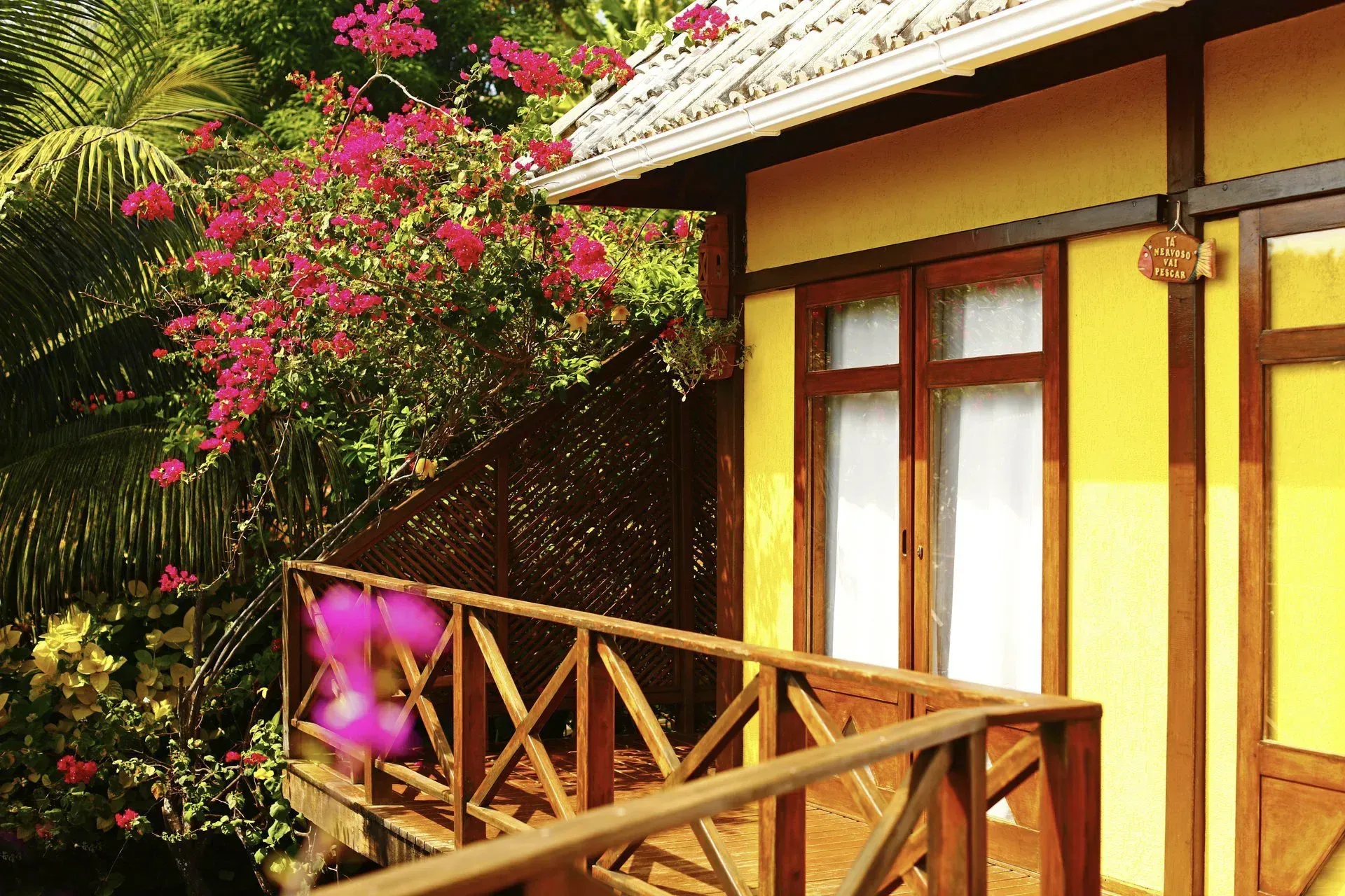 Uma casa amarela com deck de madeira e flores ao fundo