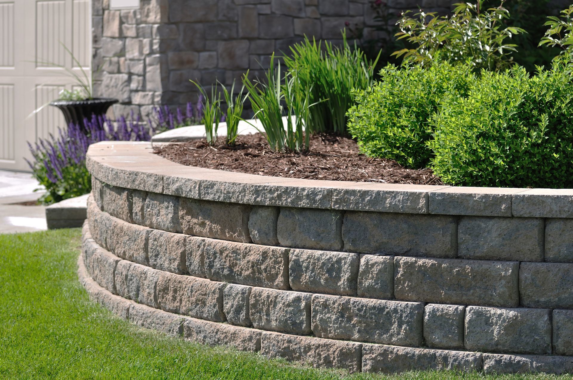 A curved, stacked stone retaining wall filled with mulch and green shrubs in a residential yard.