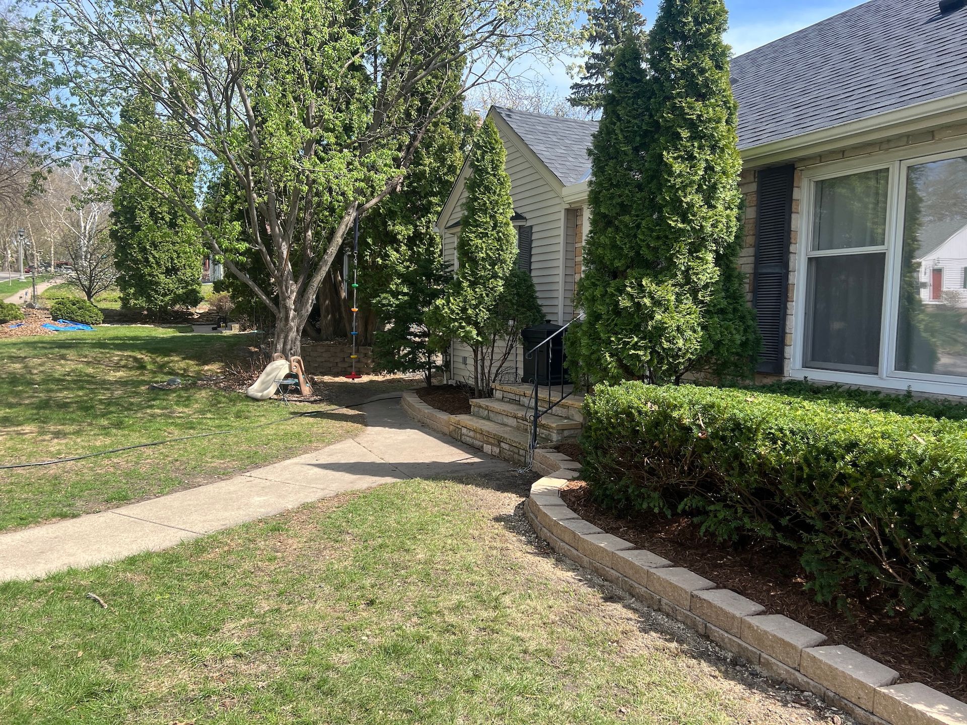 A concrete path leads to the front entrance of a light-colored house with trimmed hedges, evergreen trees, and lawn.