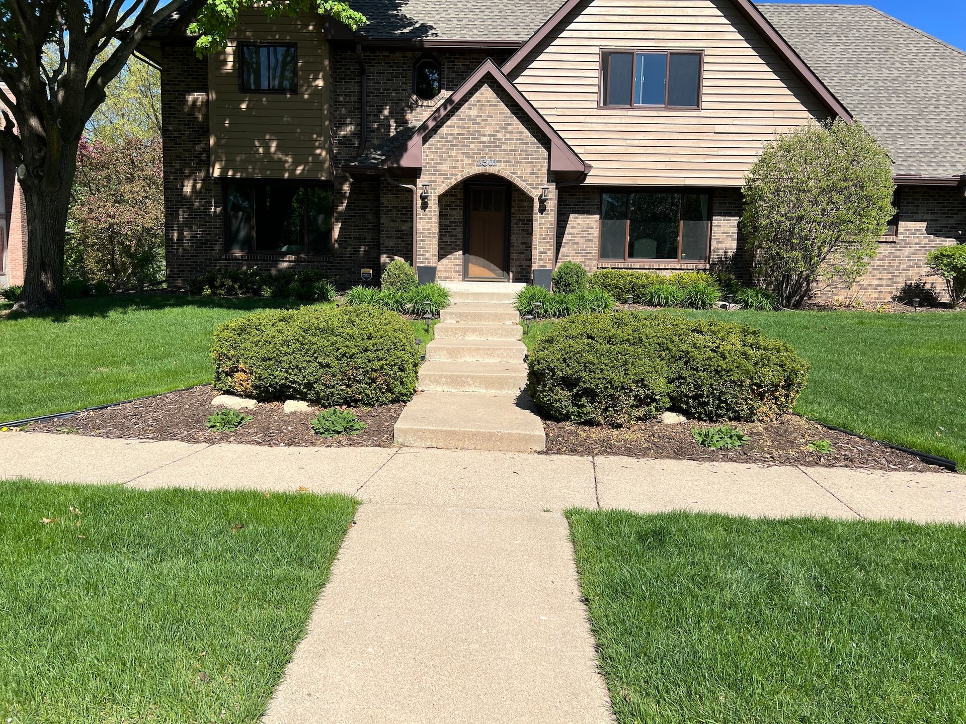 A front-facing view of a two-story brick and wood-sided house with a central concrete walkway and landscaped front yard.