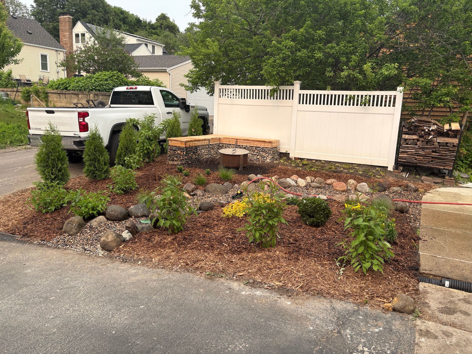 A white fence borders a garden with small evergreens, rocks, mulch, and flowers, near a parked white pickup truck.