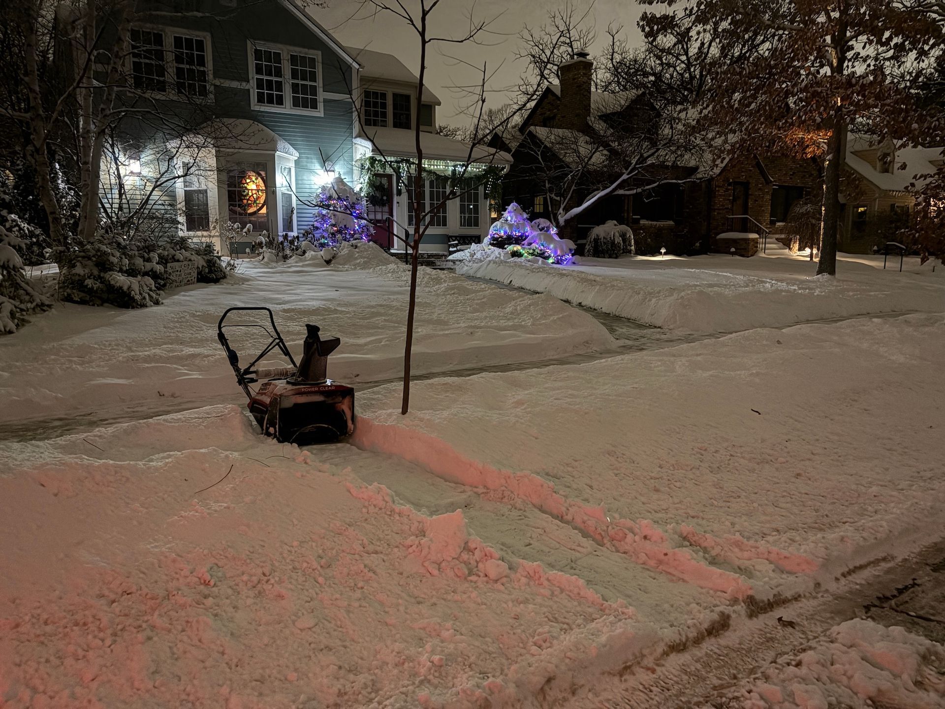 A snowblower sits on a snow-covered sidewalk at night, with a streak of pink-tinted snow leading away from it.