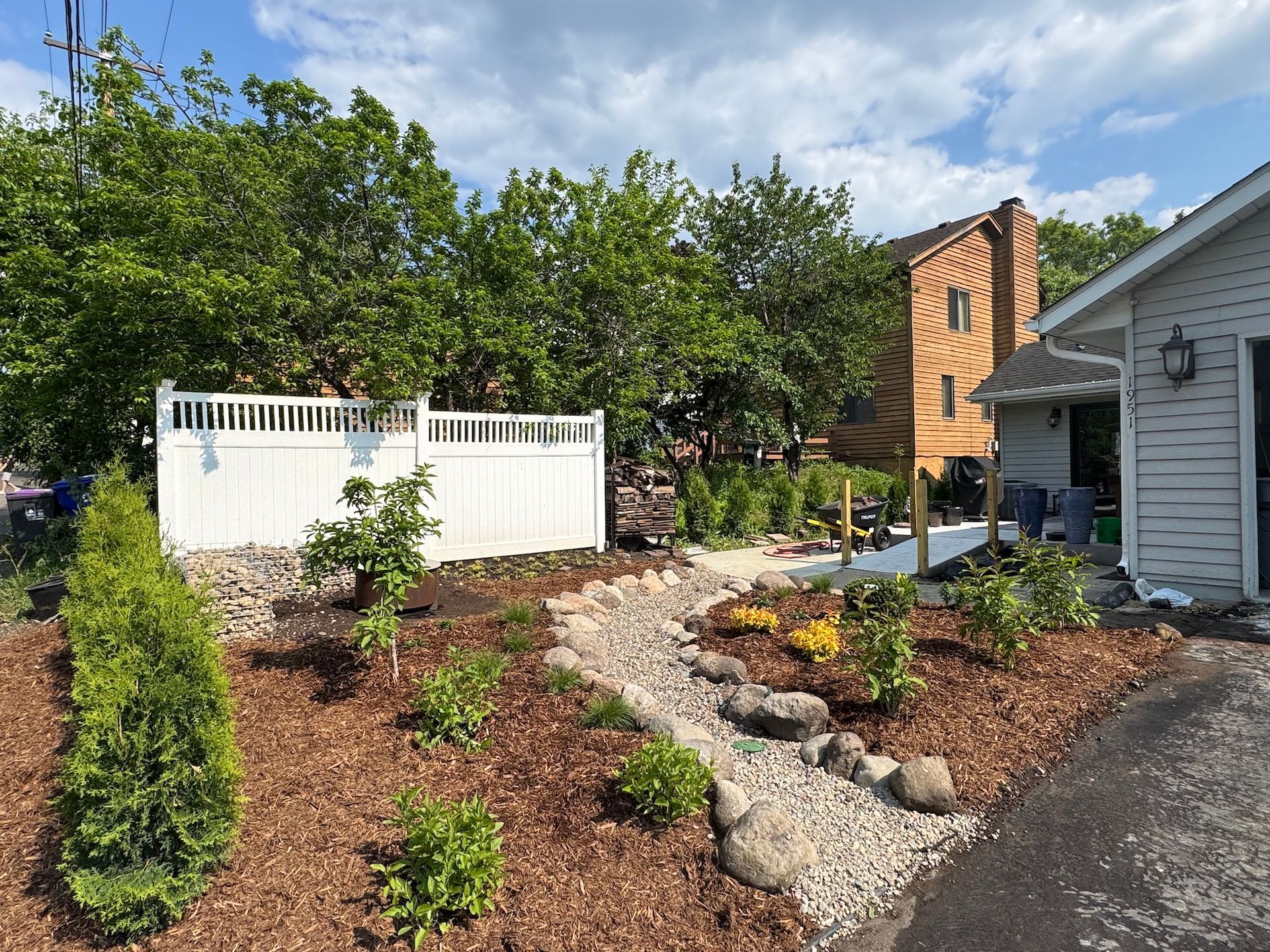 A landscaped yard features a gravel dry creek bed, brown mulch, young shrubs, and a white fence next to a house.