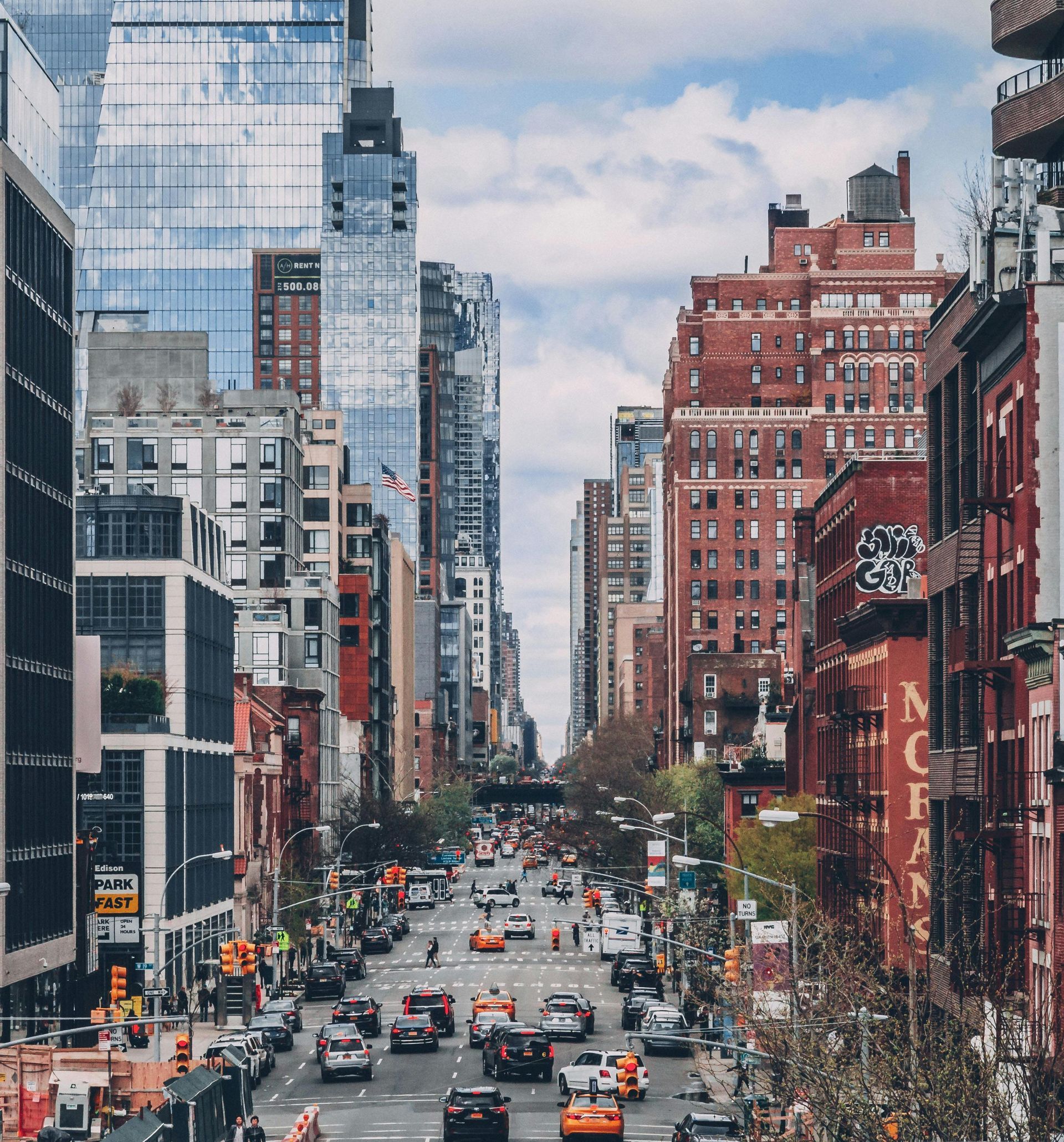 City street with tall buildings and traffic under a cloudy sky.