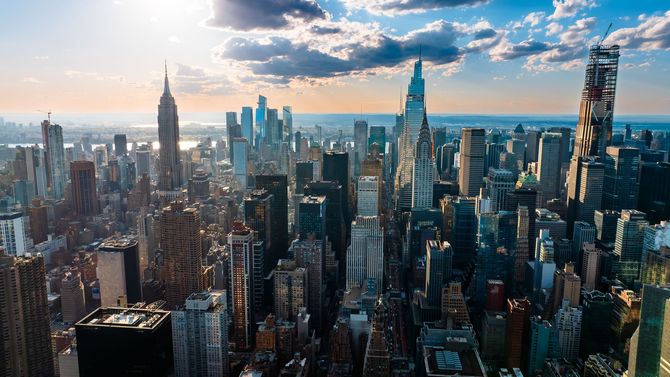 Aerial view of New York City skyline at sunset. Skyscrapers, blue sky, clouds, and a distant ocean horizon.