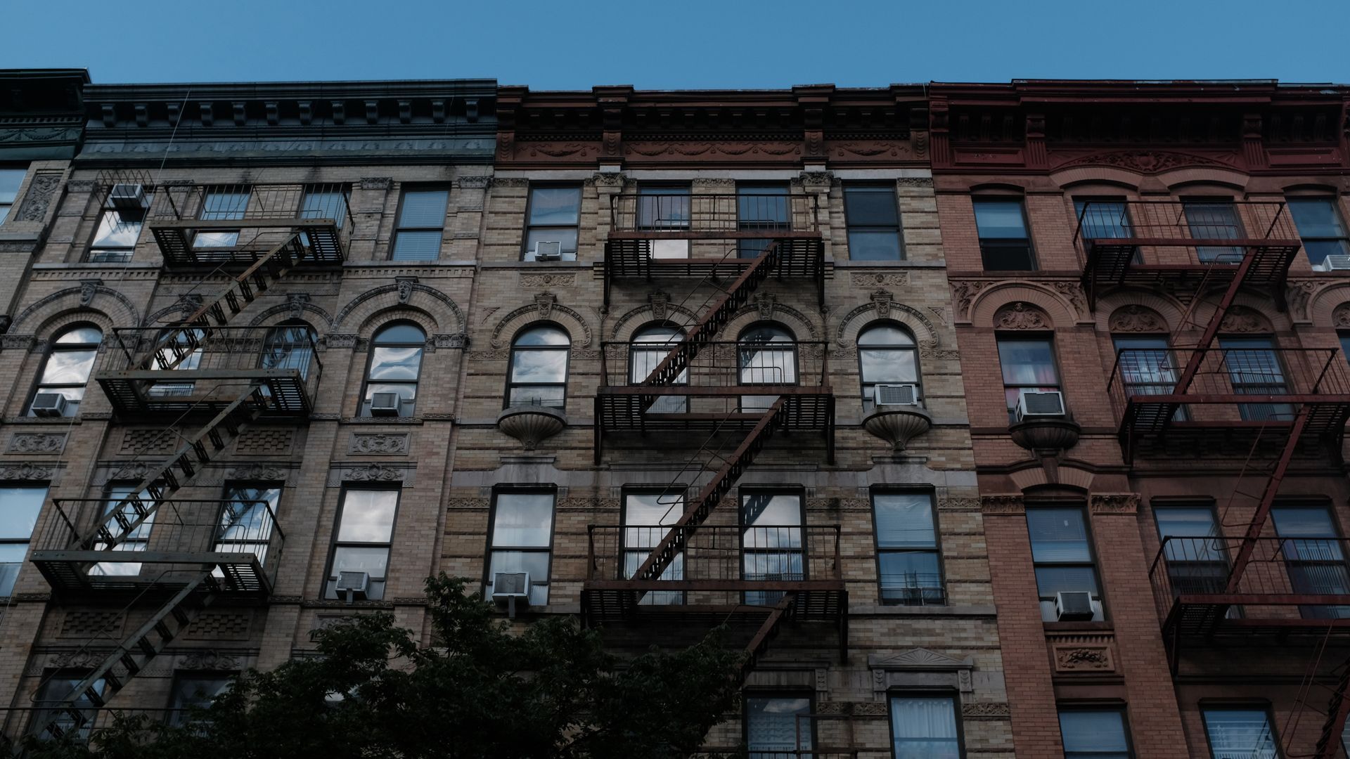 Row of old brick buildings with fire escapes and windows against a blue sky.
