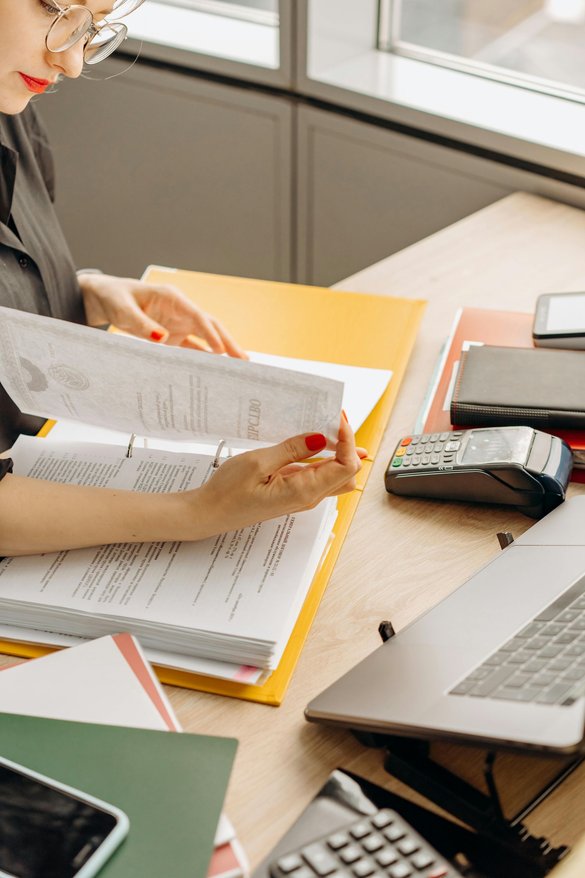 Woman examining documents at a desk with a laptop, calculator, and notepad.