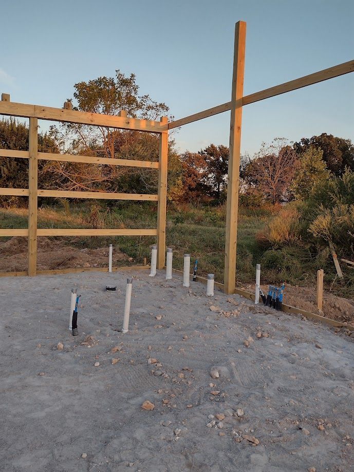 Construction site: wooden framework, plumbing pipes on a concrete slab, outdoor setting.