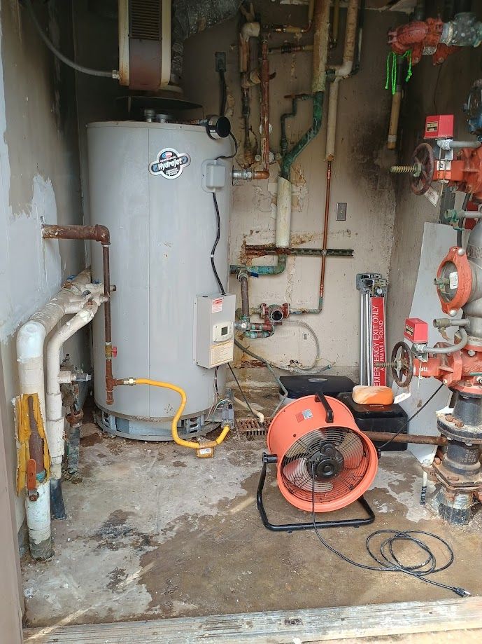 Water heater and pipes in a utility room, with a fan on the floor.