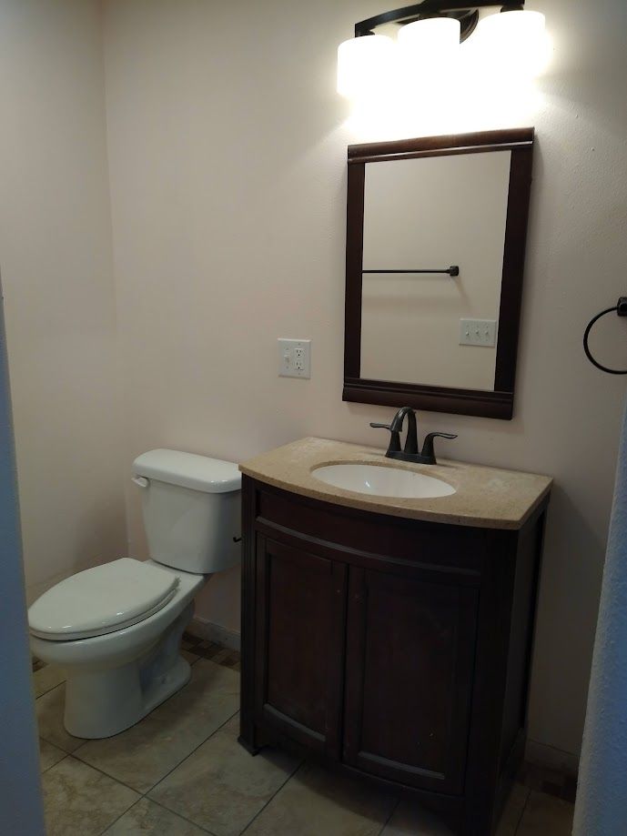 Small bathroom with toilet, vanity, mirror, and light fixture.  Beige walls and tile floor. Dark brown vanity.