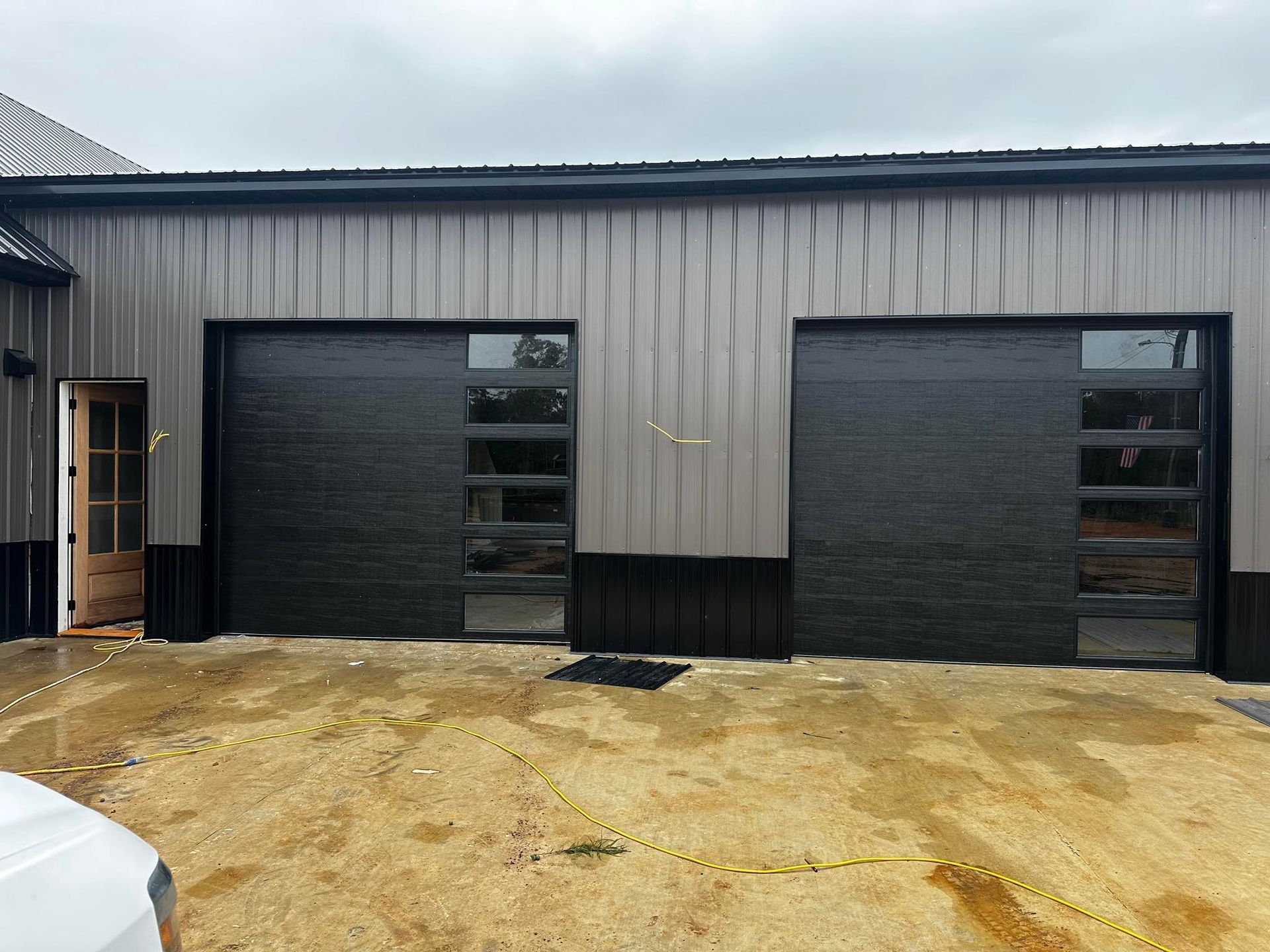 Two black garage doors with glass panels on a building with light gray siding and a wooden door.