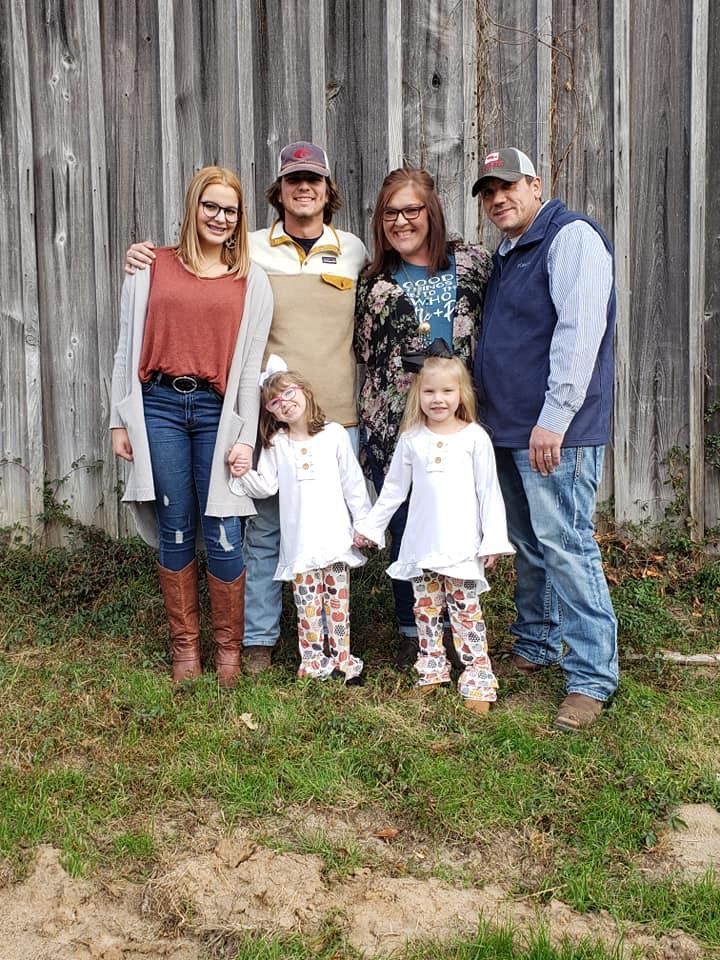 Family of six poses in front of a weathered wooden wall. Two children hold hands.