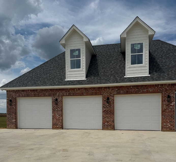 A red brick detached three-car garage with white doors, a dark shingled roof, and two white dormers under a cloudy sky. A red brick detached three-car garage with white doors, a dark shingled roof, and two white dormers under a cloudy sky.