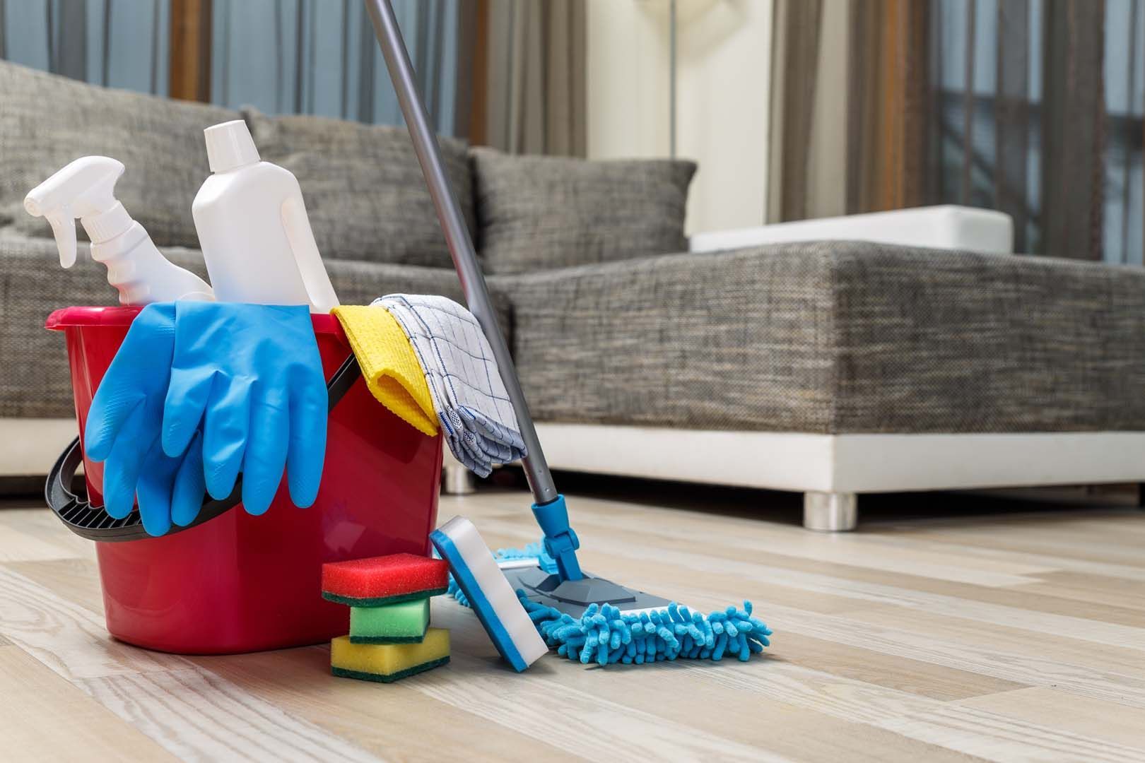 A red bucket filled with cleaning supplies, including gloves, bottles, and sponges, sits next to a mop on a wooden floor.