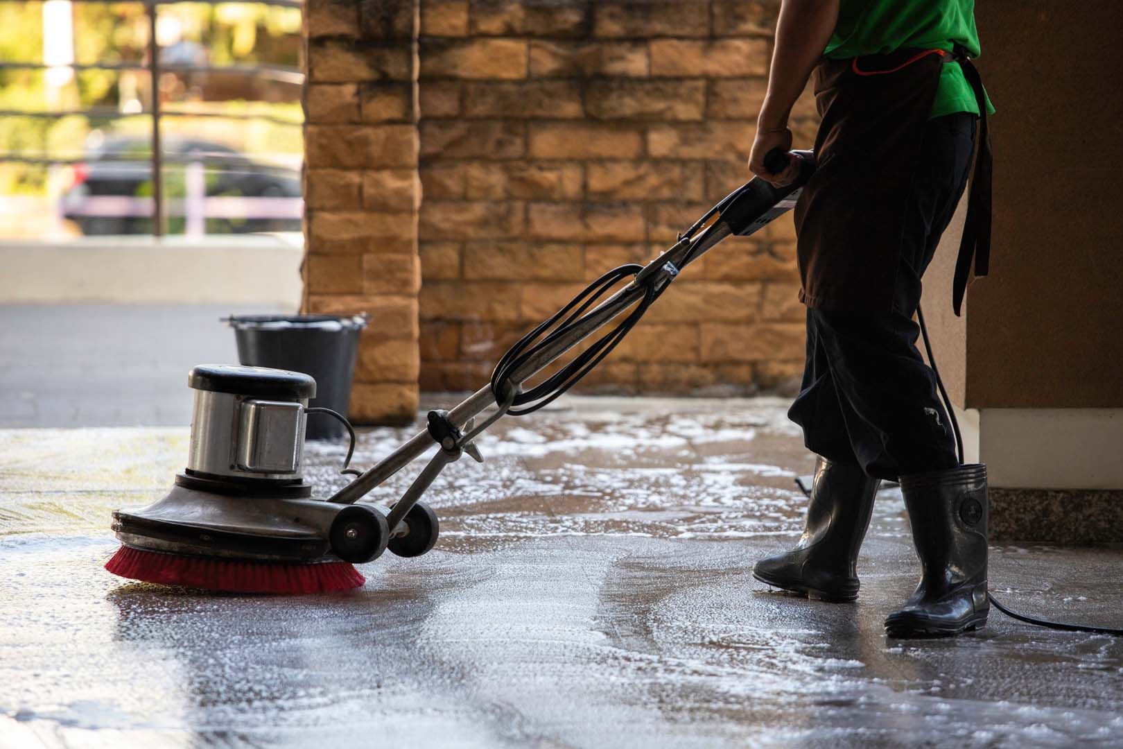 A person in a green shirt and dark apron uses a floor buffer to scrub a wet, soapy floor.