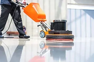 A person uses an orange floor polishing machine to buff and clean a shiny, reflective indoor floor.