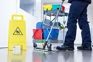 A worker in dark blue uniform mops a shiny floor next to a yellow caution sign and a rolling cleaning cart with buckets.