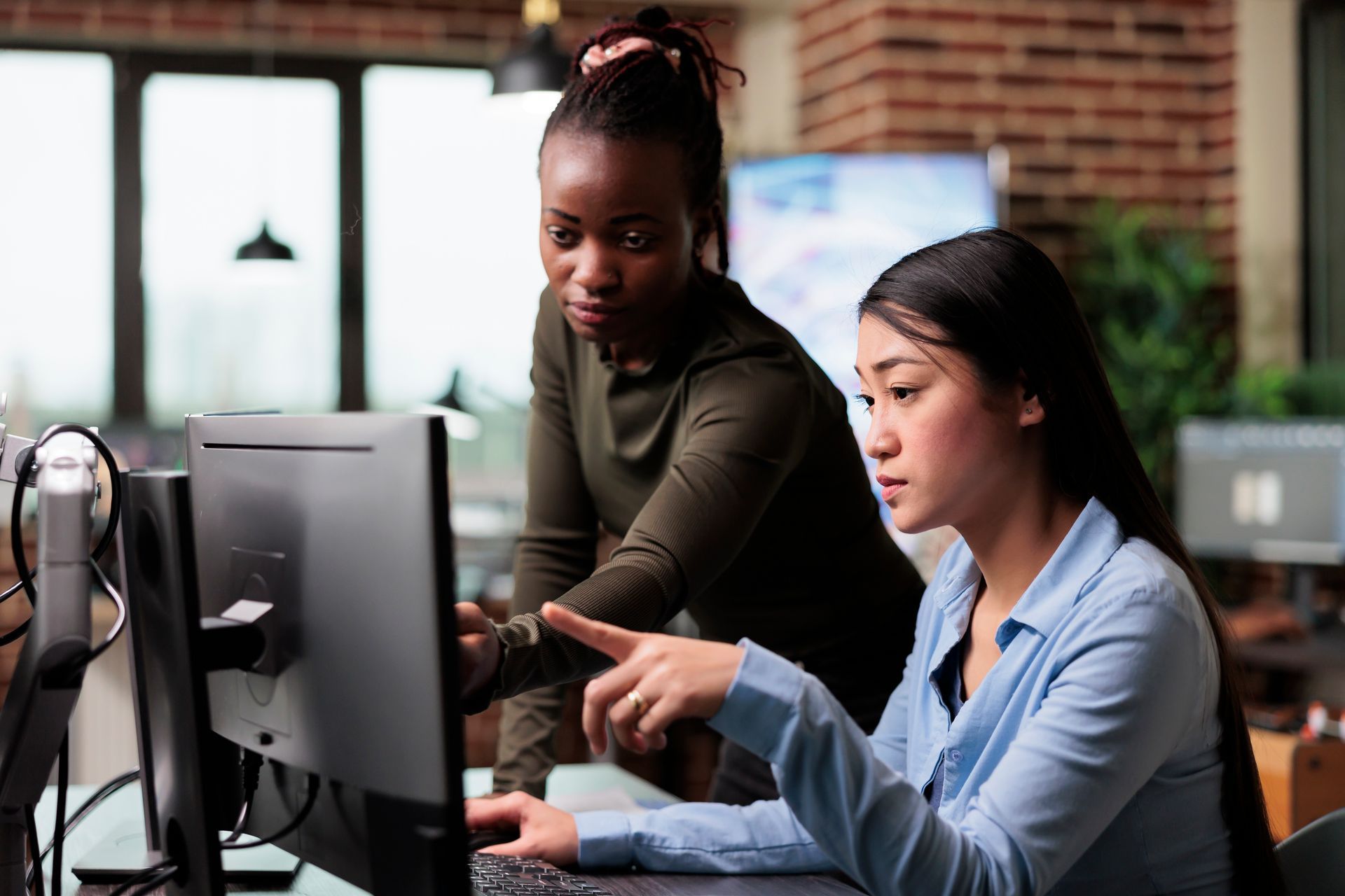 Two women working on a computer together in an office, one points at the screen.