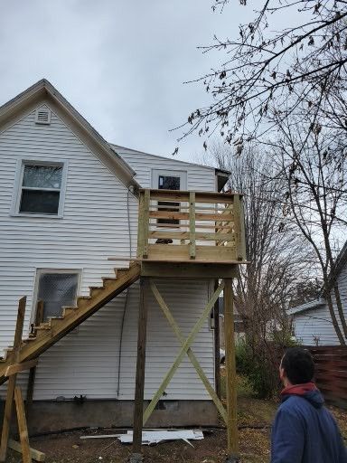 A man is standing in front of a house with a wooden deck and stairs.