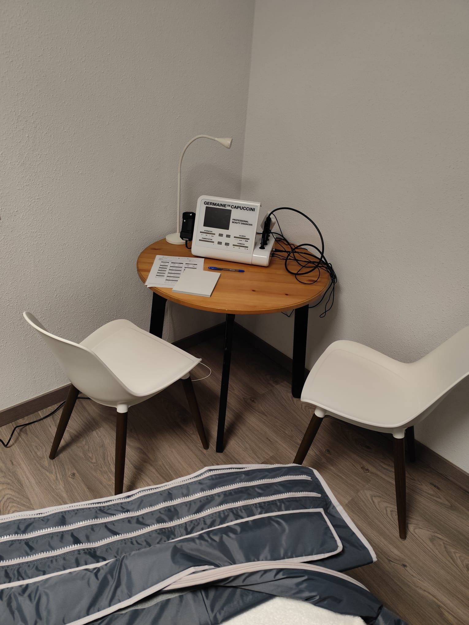 Una pequeña mesa redonda de madera con equipo médico y dos sillas blancas en un rincón. Una almohadilla térmica terapéutica está en el suelo.