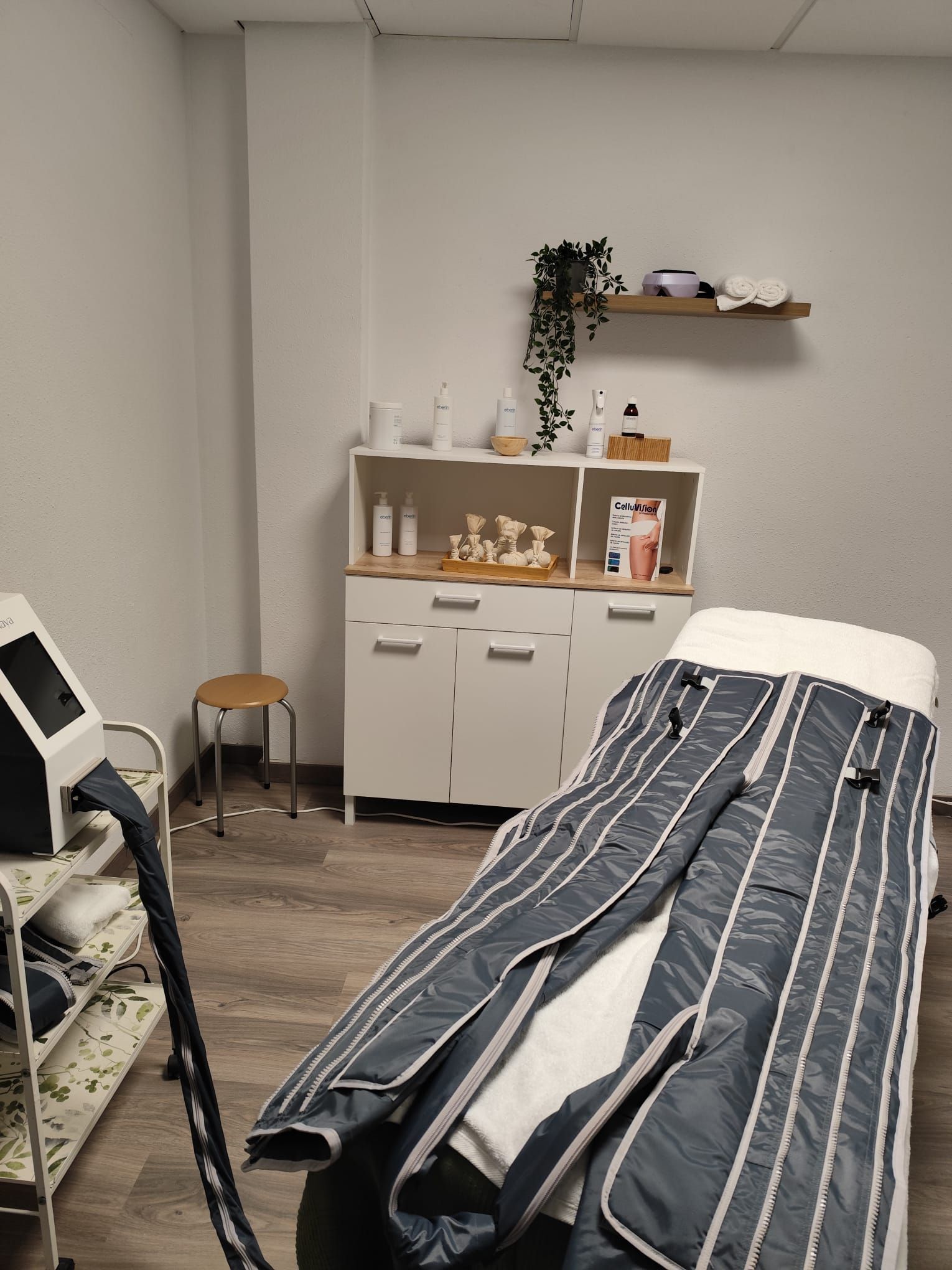 Una sala de tratamientos de spa con camilla y equipo de terapia. Un armario contiene productos de belleza y una estantería muestra la decoración.