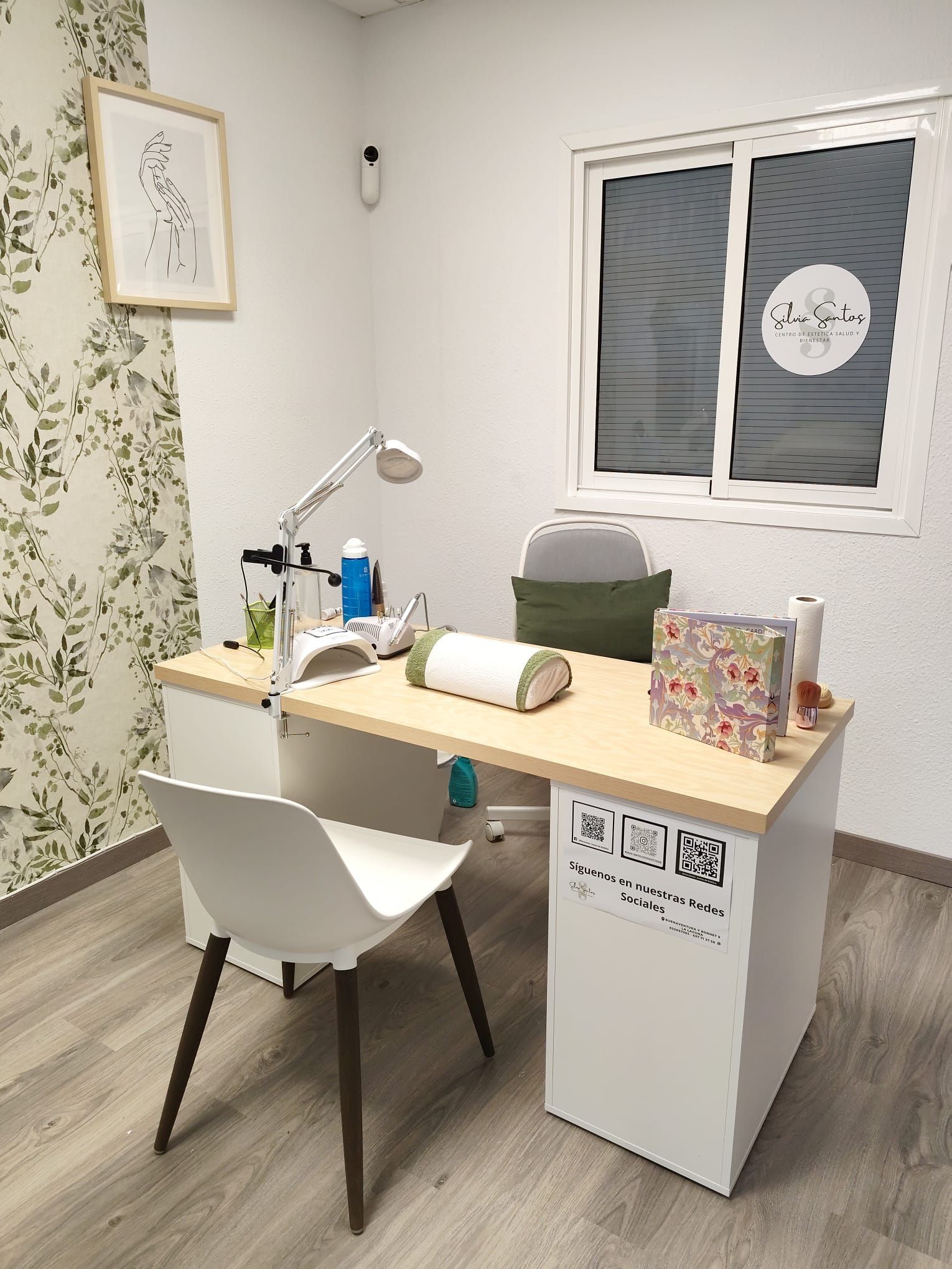 Una estación de trabajo de salón de uñas con un escritorio blanco, una silla y una ventana con diseño esmerilado. También incluye un papel pintado decorativo con diseño de hojas.