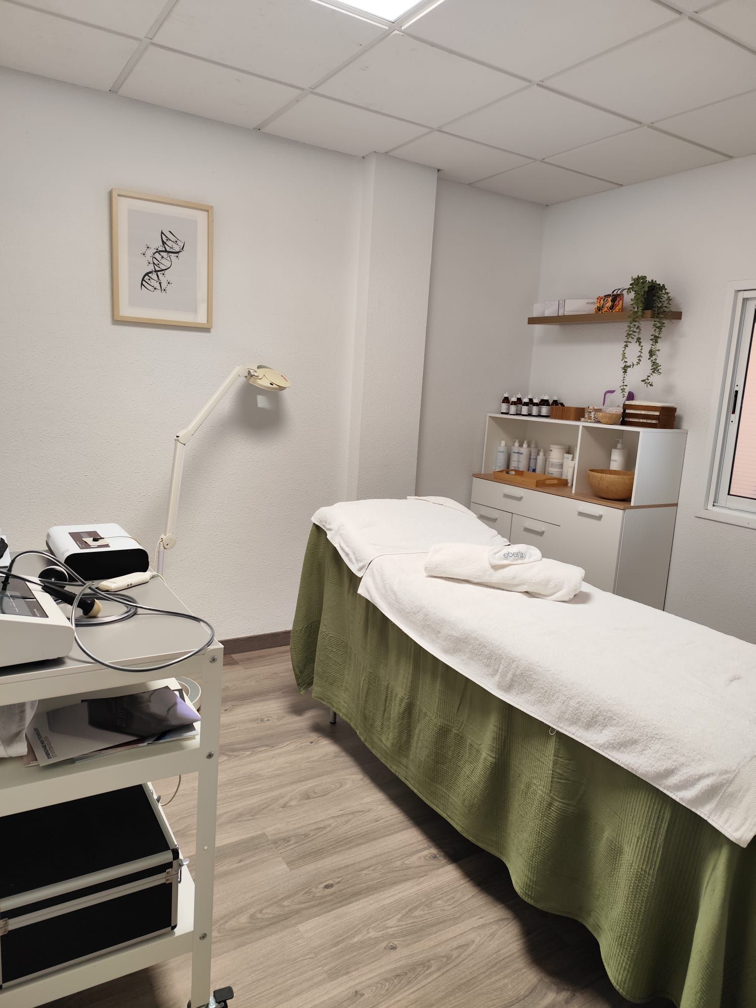 Una sala de tratamientos de spa con una camilla de masajes cubierta con sábanas blancas y una manta verde. Hay un carrito médico con equipo a la izquierda y un armario con productos a la derecha.