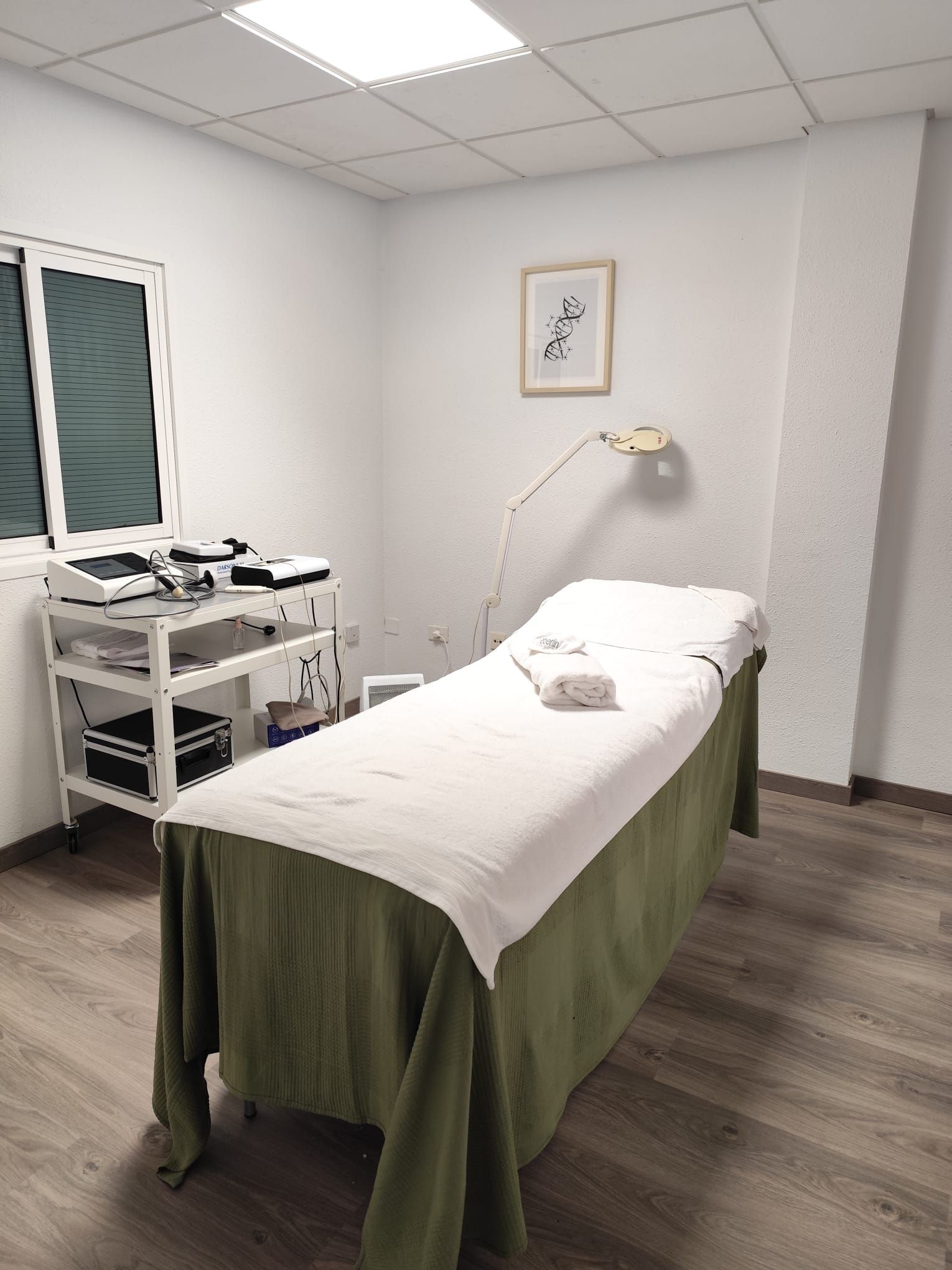 Una sala de masajes con una mesa de masajes cubierta con tela blanca y verde, equipo médico y una obra de arte enmarcada.